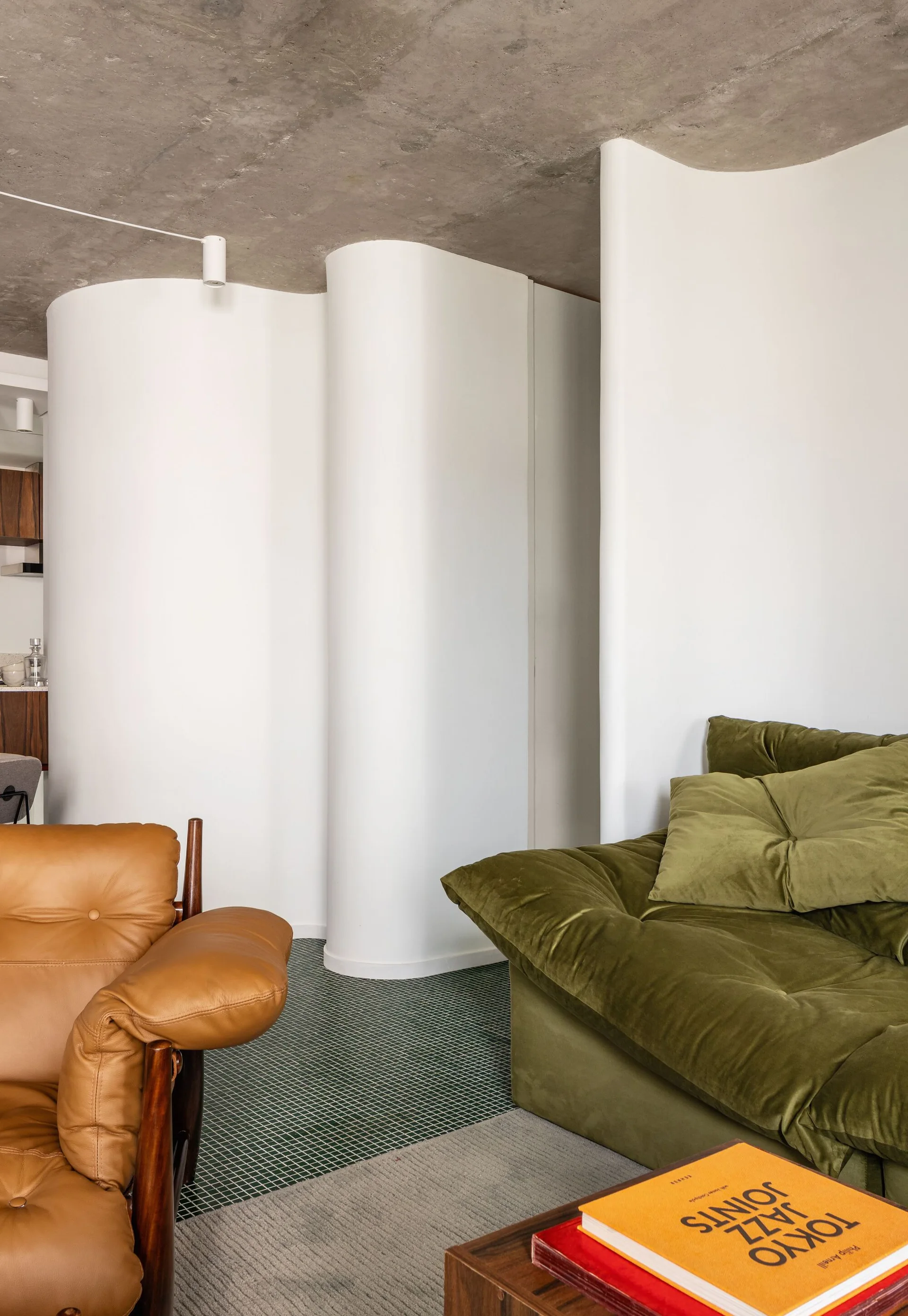 An olive green velvet sofa and tan leather armchair set against white curved partition walls under a textured concrete ceiling.