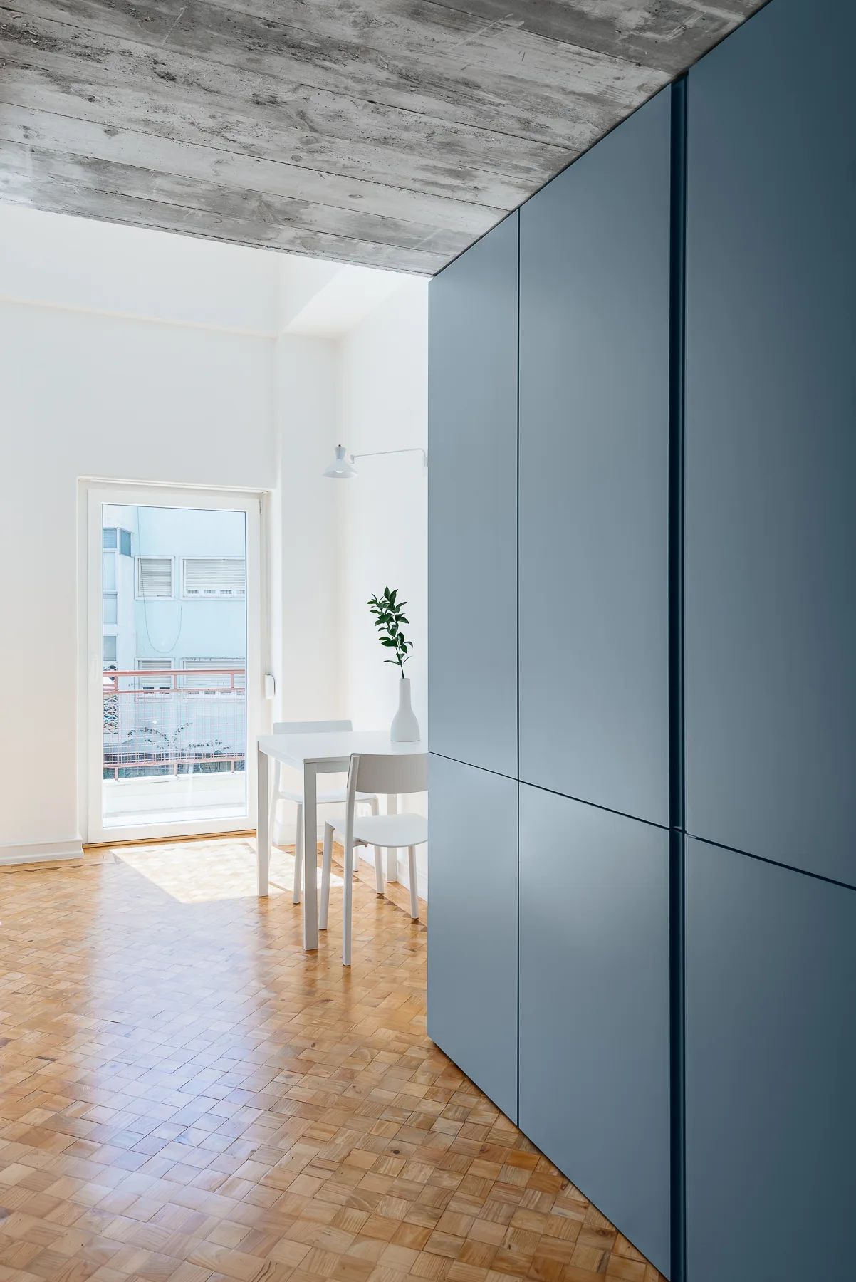 A minimalist dining room features a large matte blue cabinet unit, an exposed concrete ceiling, a white dining set, and parquet wood flooring.