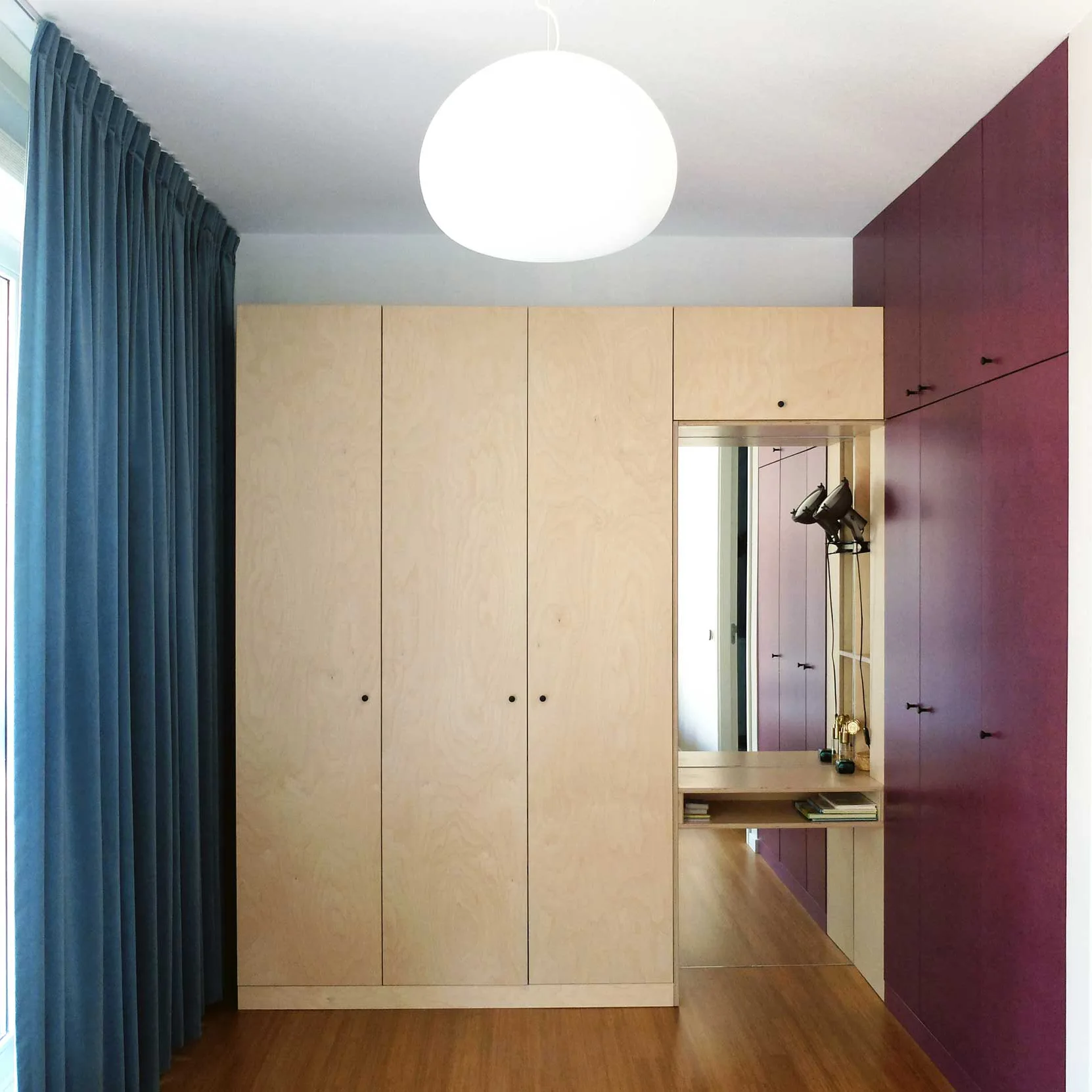 A plywood wardrobe stands next to deep purple cabinetry with an integrated vanity niche, a blue curtain, and a globe ceiling light.