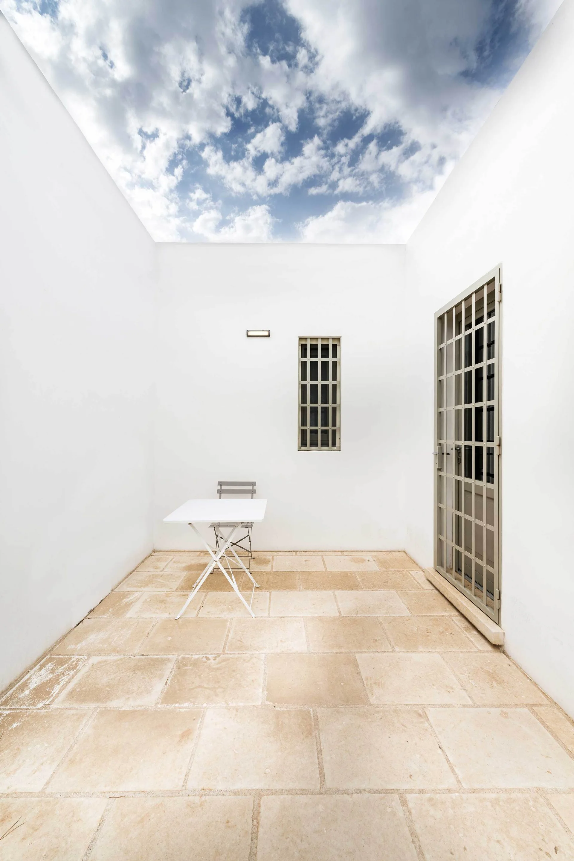 A minimalist white walled courtyard features stone flagstone flooring, a small folding table and chair set, and a metal-grille door.
