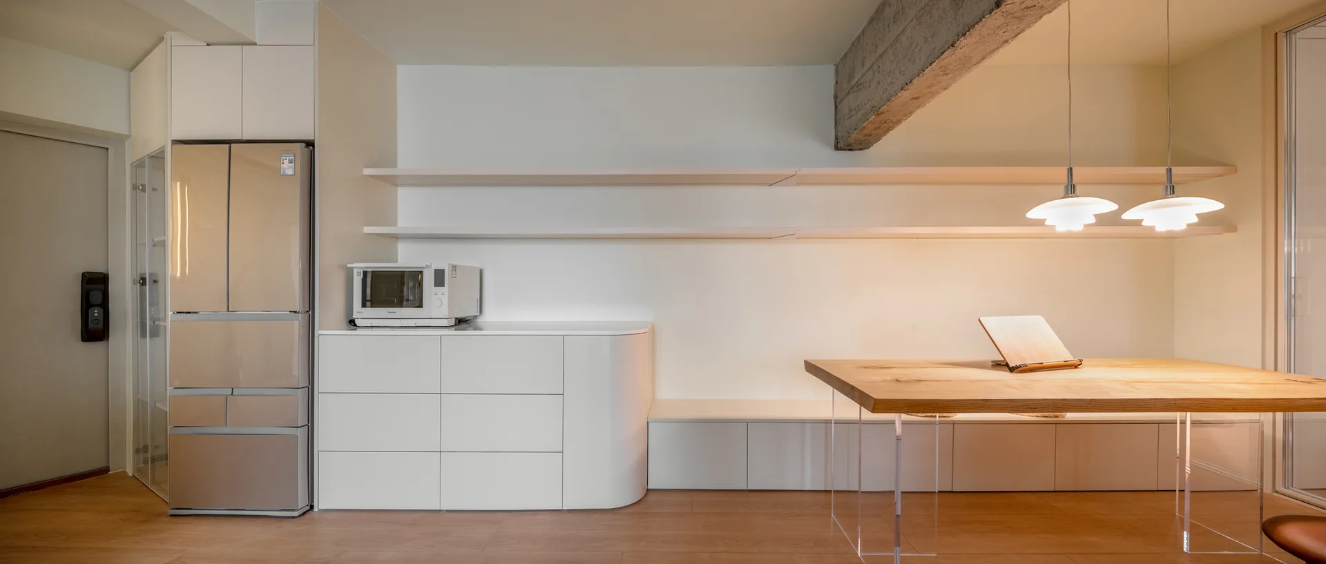 A minimalist kitchen and dining area features a wood table with clear acrylic legs, low white cabinets, and an exposed concrete ceiling beam.