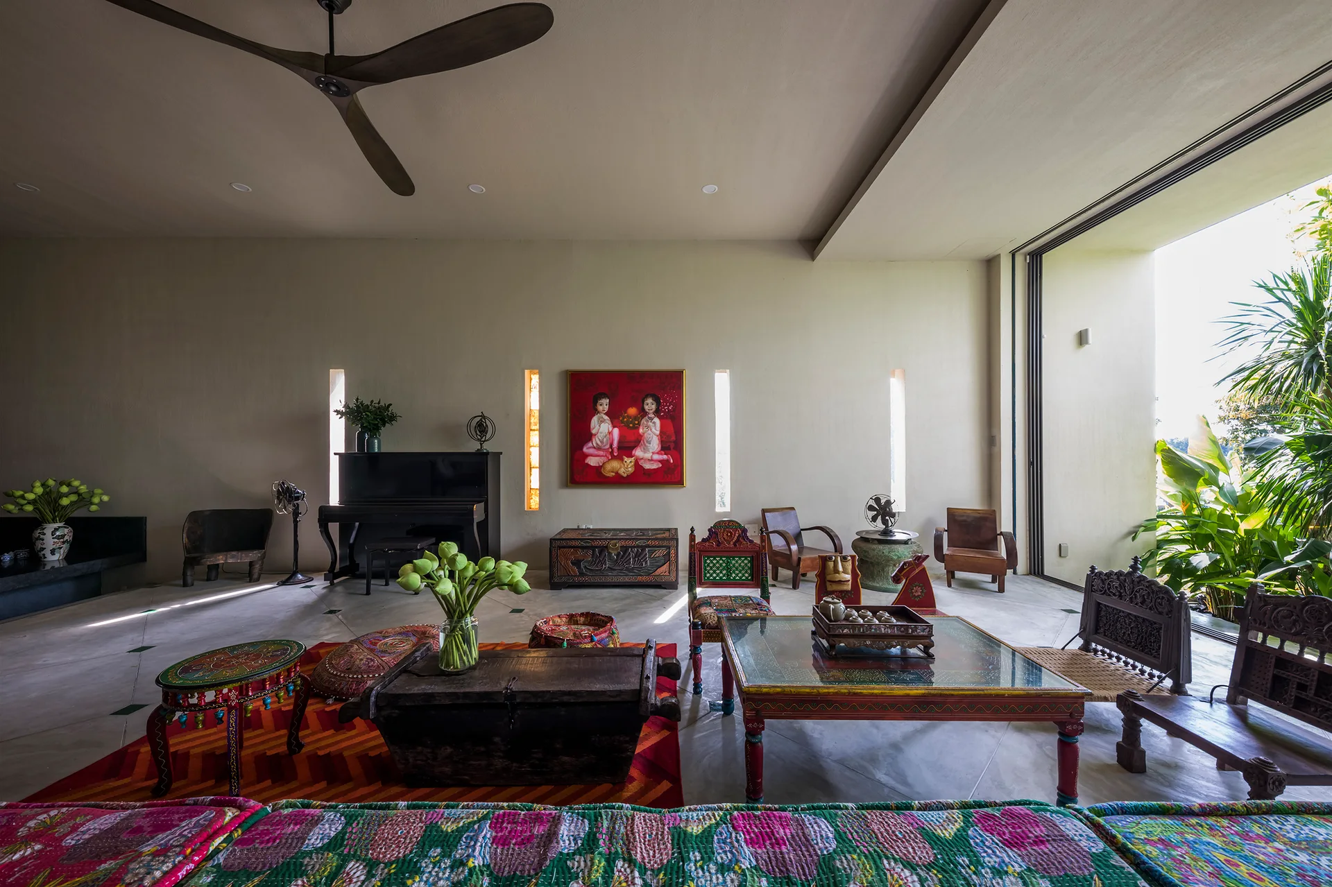 An eclectic living room features a low carved wood coffee table, colorful patterned textiles, a black piano, and tall narrow wall niches.