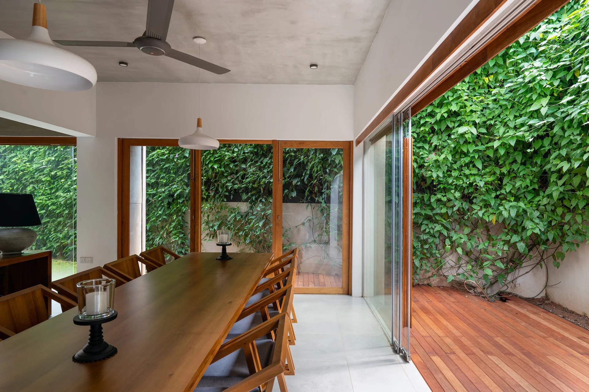 A wooden dining table and chairs in a dining room with large sliding glass doors opening to a wood deck with green climbing vines.