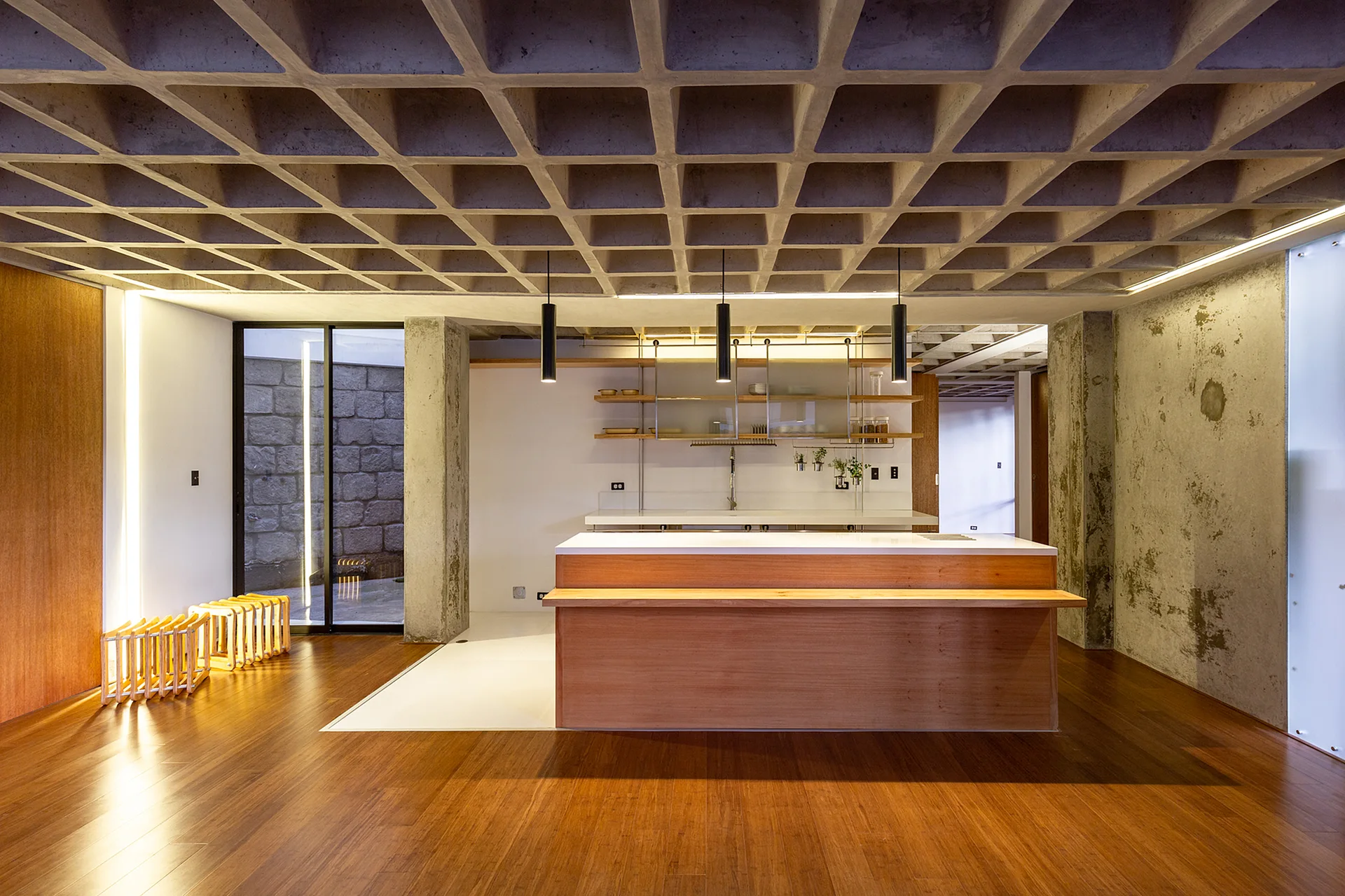 A modern kitchen featuring a wood island, cylindrical pendant lights, and an exposed waffle concrete ceiling above wood flooring.