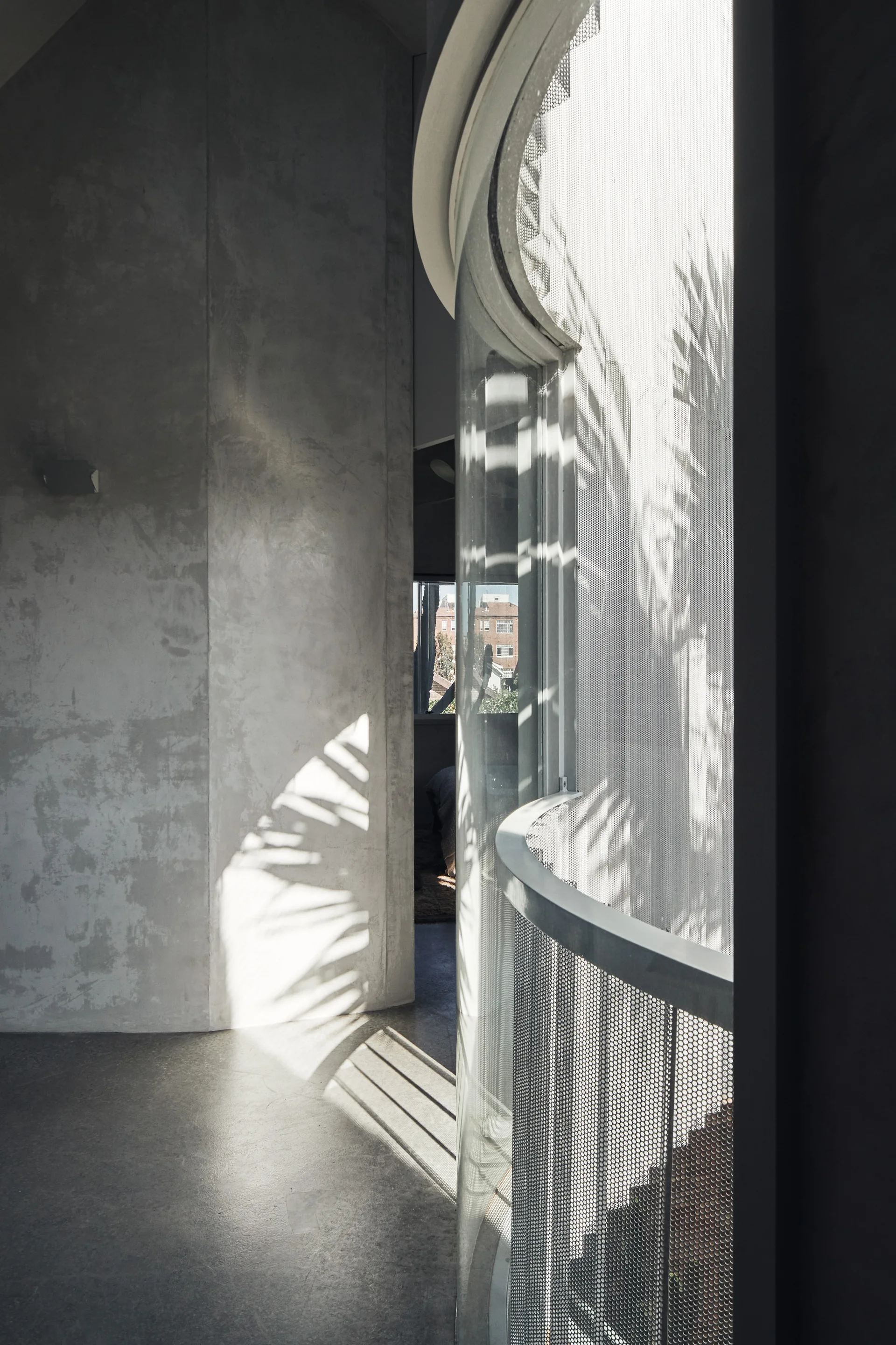 A minimalist interior hallway features a curved glass window, a perforated metal railing, and raw concrete walls illuminated by natural sunlight.