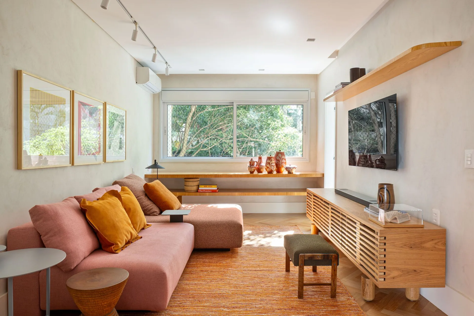 A warm living room featuring a pink sectional sofa, an orange textured rug, and a slatted wood media console beneath a floating shelf.