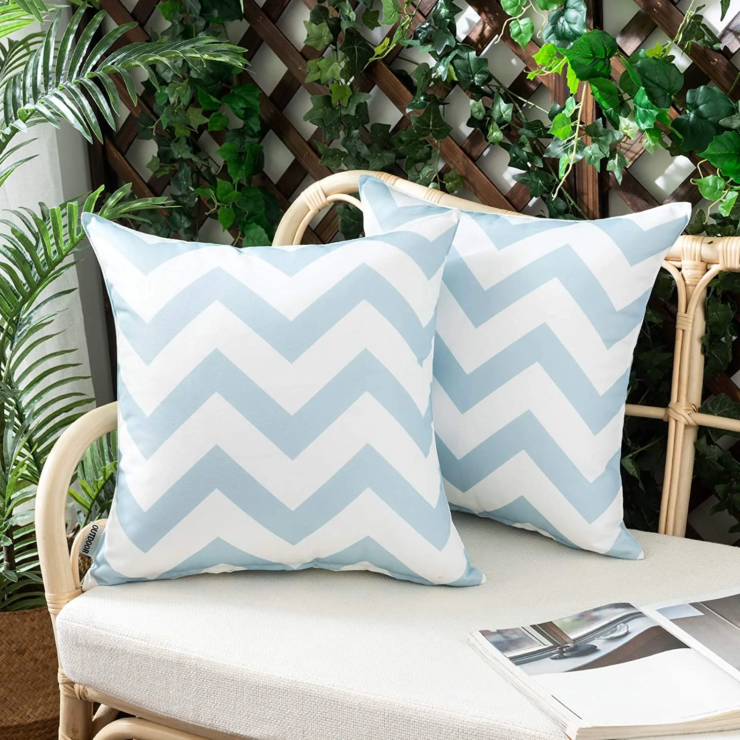 Two light blue and white chevron patterned throw pillows sit on a rattan patio bench in front of a wooden lattice trellis with green ivy.