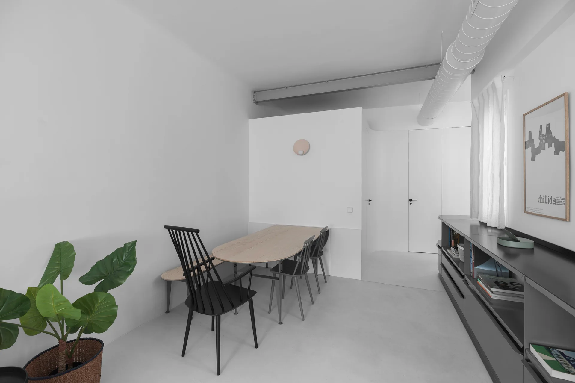 A minimalist dining room features a light wood oval table with black spindle chairs, a grey sideboard, and exposed white ceiling ductwork.