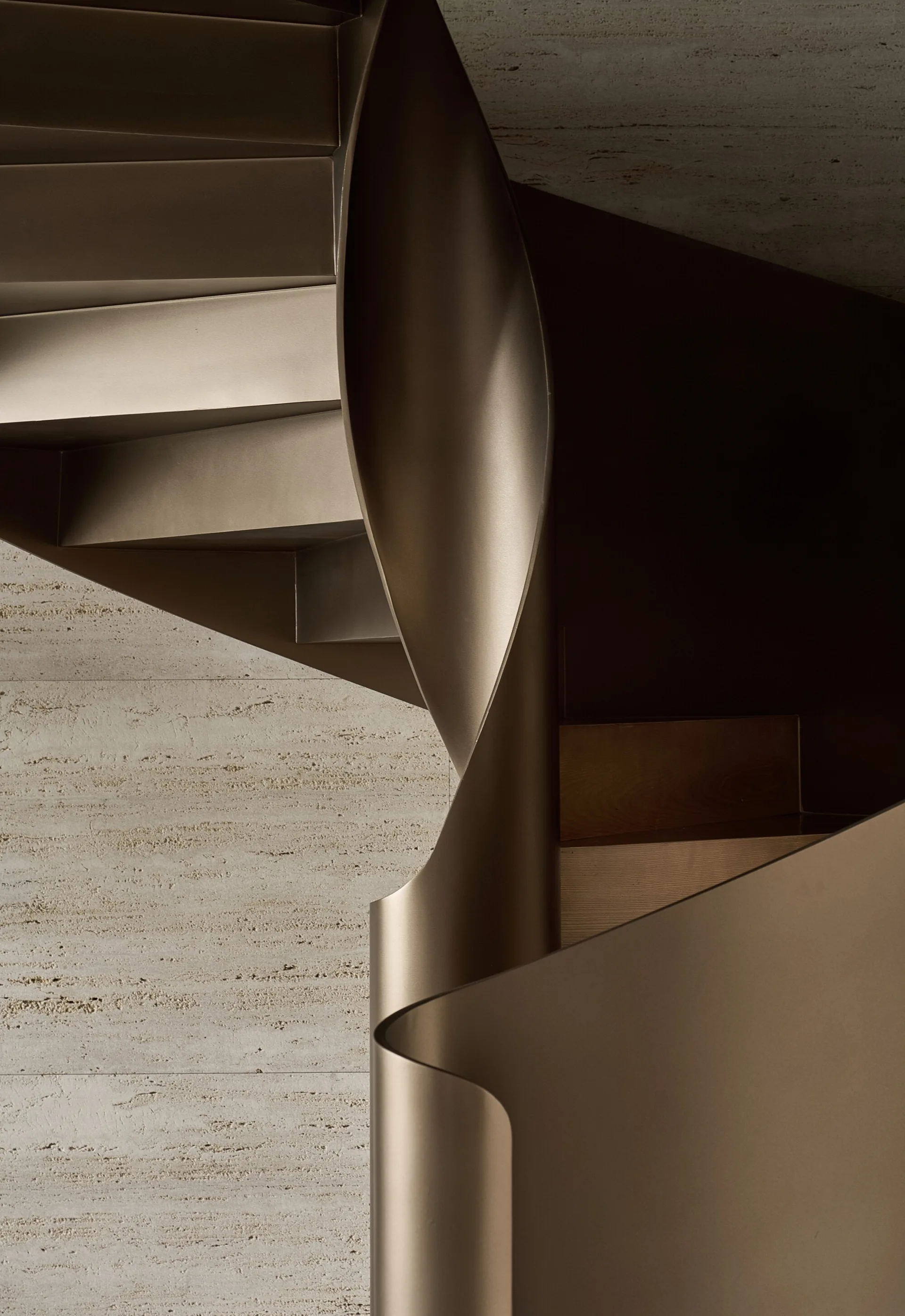 Close-up of a bronze metal spiral staircase with a curved ribbon railing set against a textured beige stone wall.