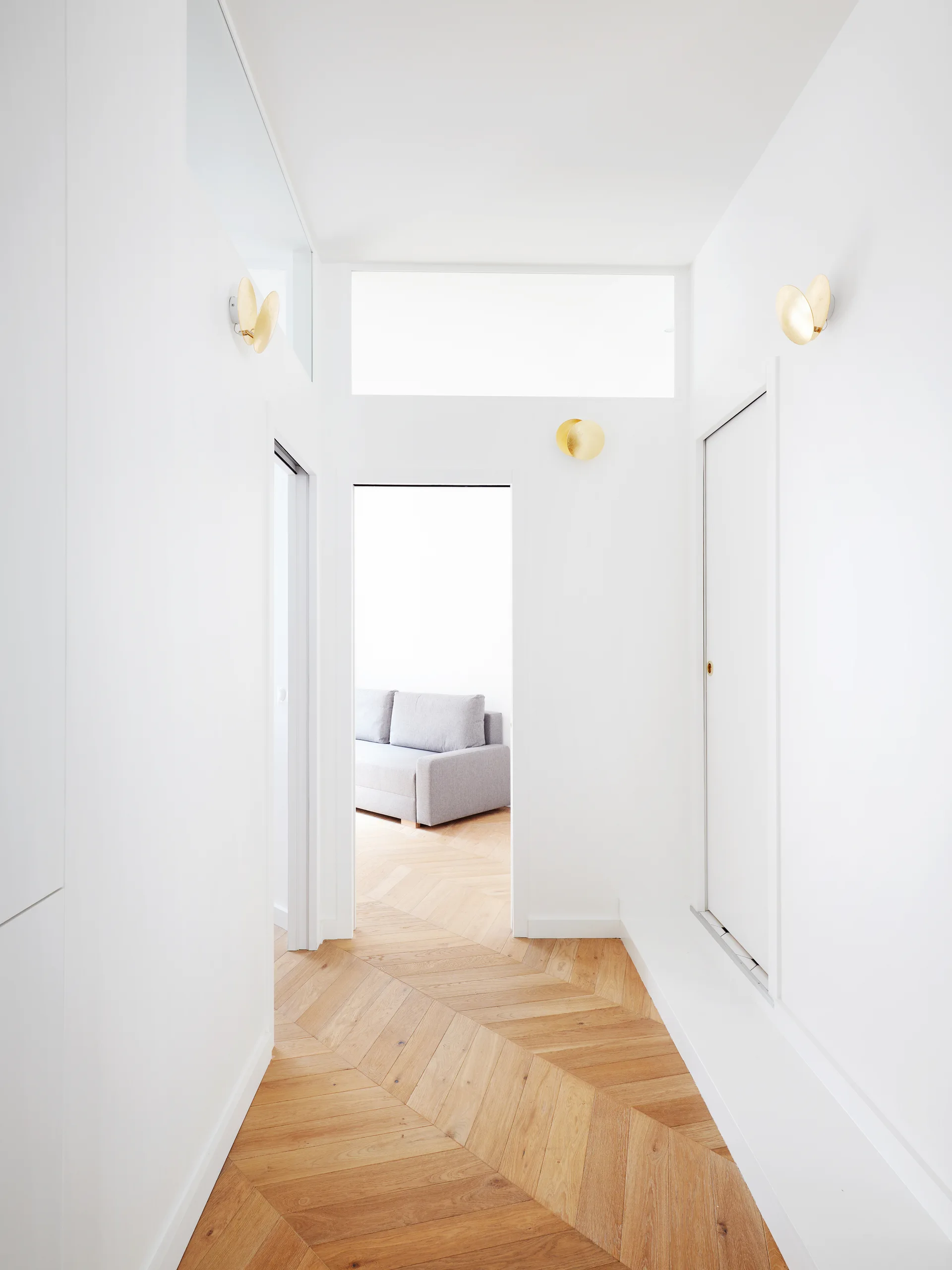A minimalist white hallway featuring light wood chevron flooring, a transom window over an open doorway, and gold circular wall sconces.