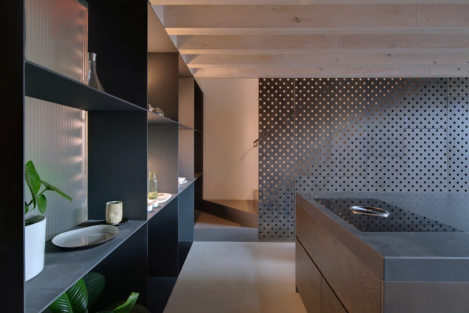 A contemporary kitchen with a dark grey island, matte black shelving, a perforated wood partition wall, and exposed light wood ceiling beams.