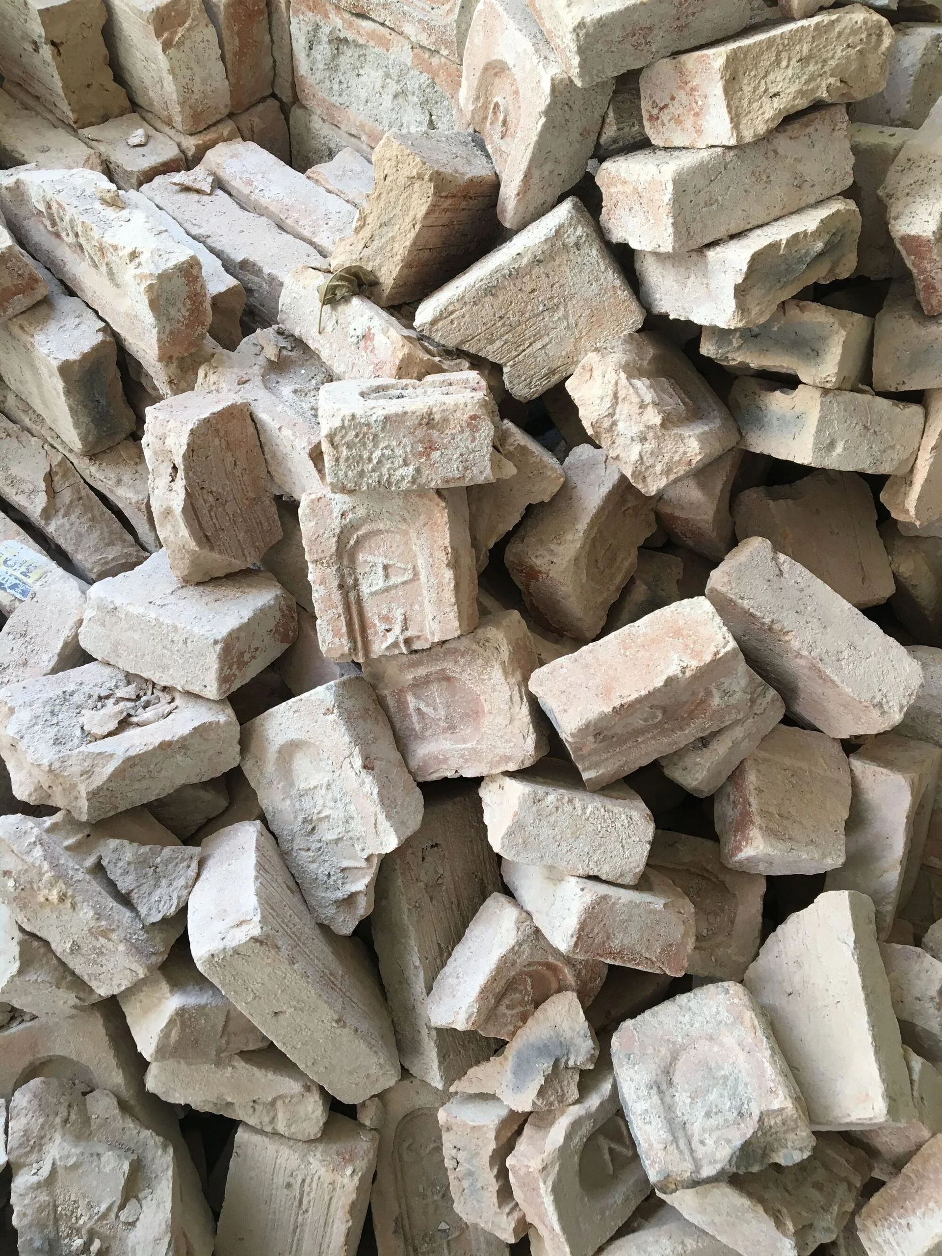 A pile of salvaged antique clay bricks in light beige and tan shades shows textured surfaces and embossed vintage manufacturer stamps.