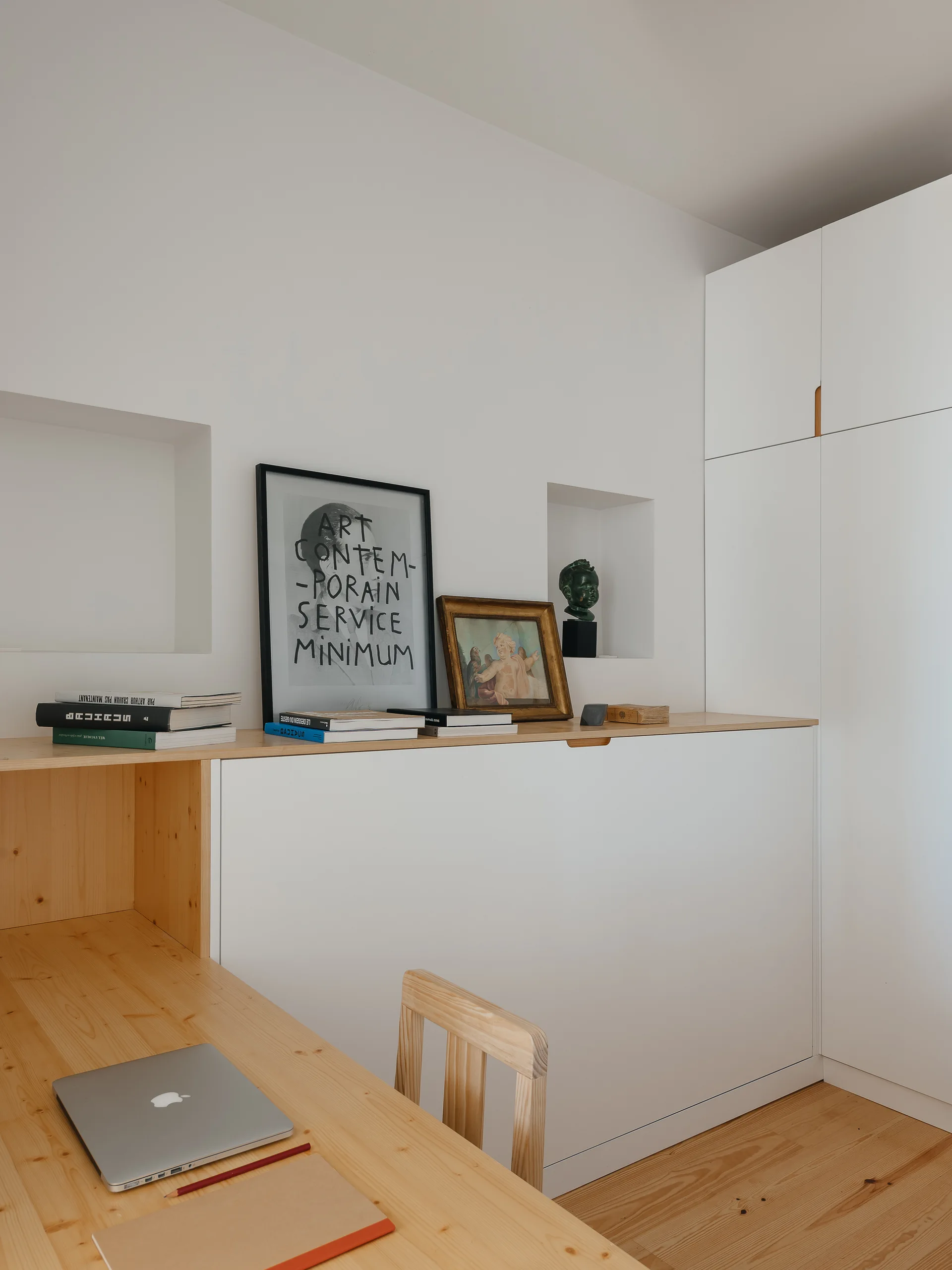 A minimalist home office featuring a light wood desk, a wooden chair, and matte white built-in storage cabinets against a plain wall with recessed niches.