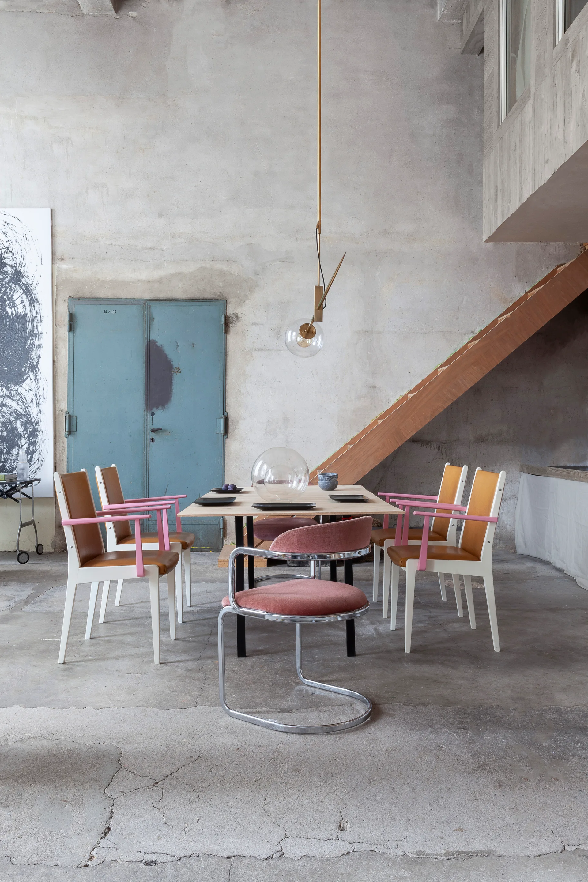 A modern wood dining table and chairs with pink accents set against raw concrete walls and a wooden staircase in an industrial loft.