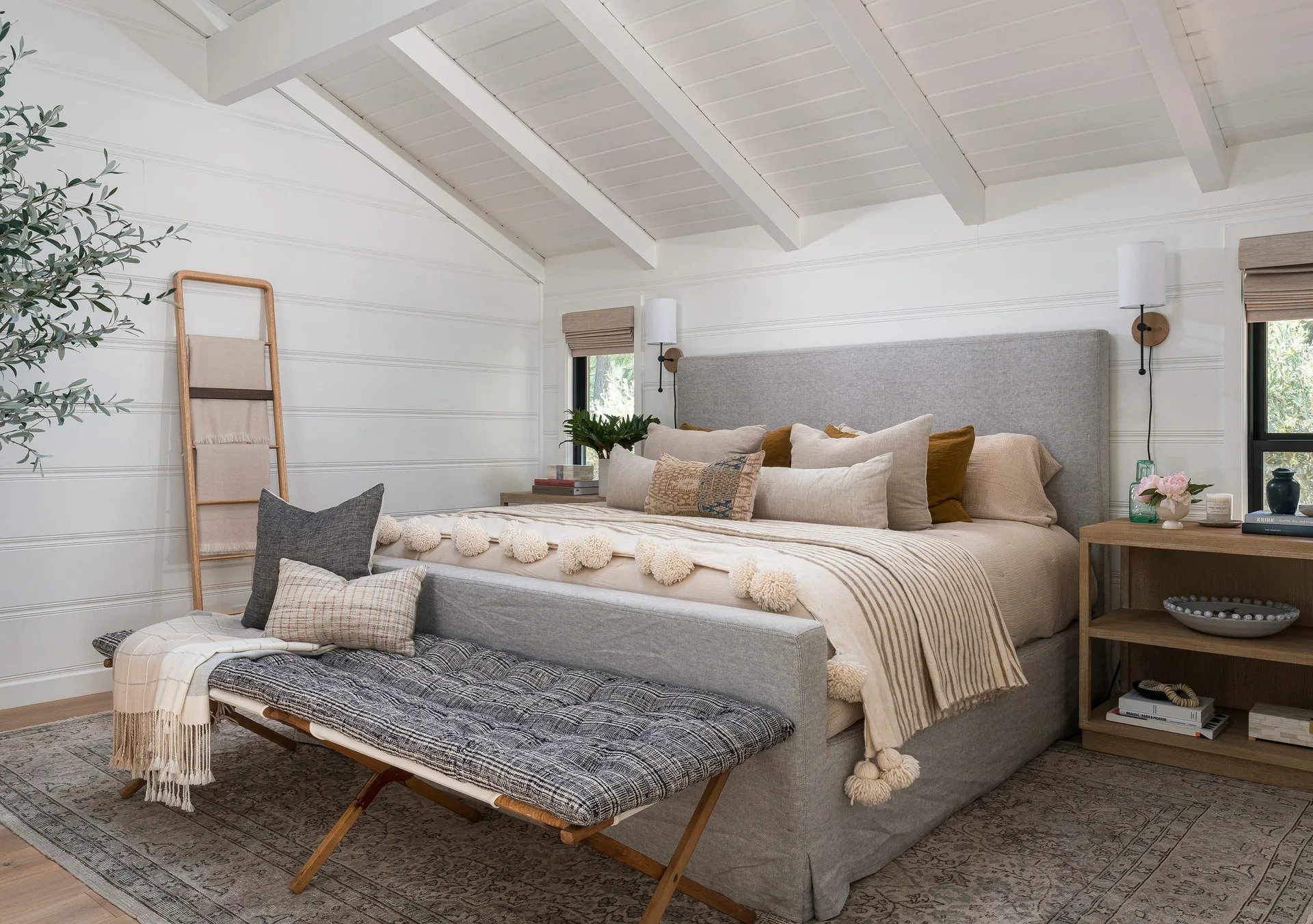 A white bedroom features a grey upholstered bed, a vaulted ceiling with beams, horizontal wall paneling, and a patterned bench at the foot of the bed.
