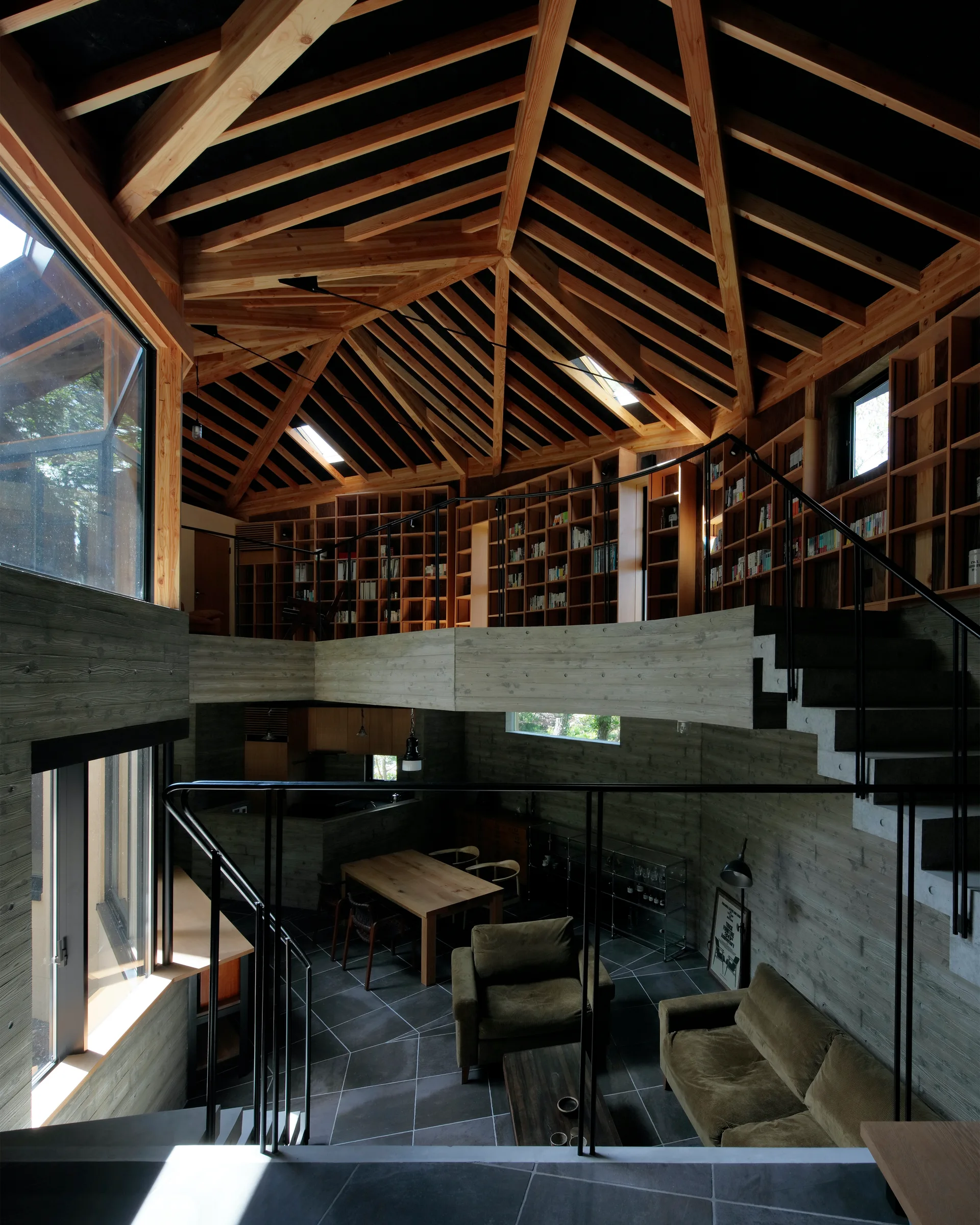 A double-height concrete living room featuring olive green seating, a dining area, and a wooden mezzanine library under an intricate exposed timber ceiling.