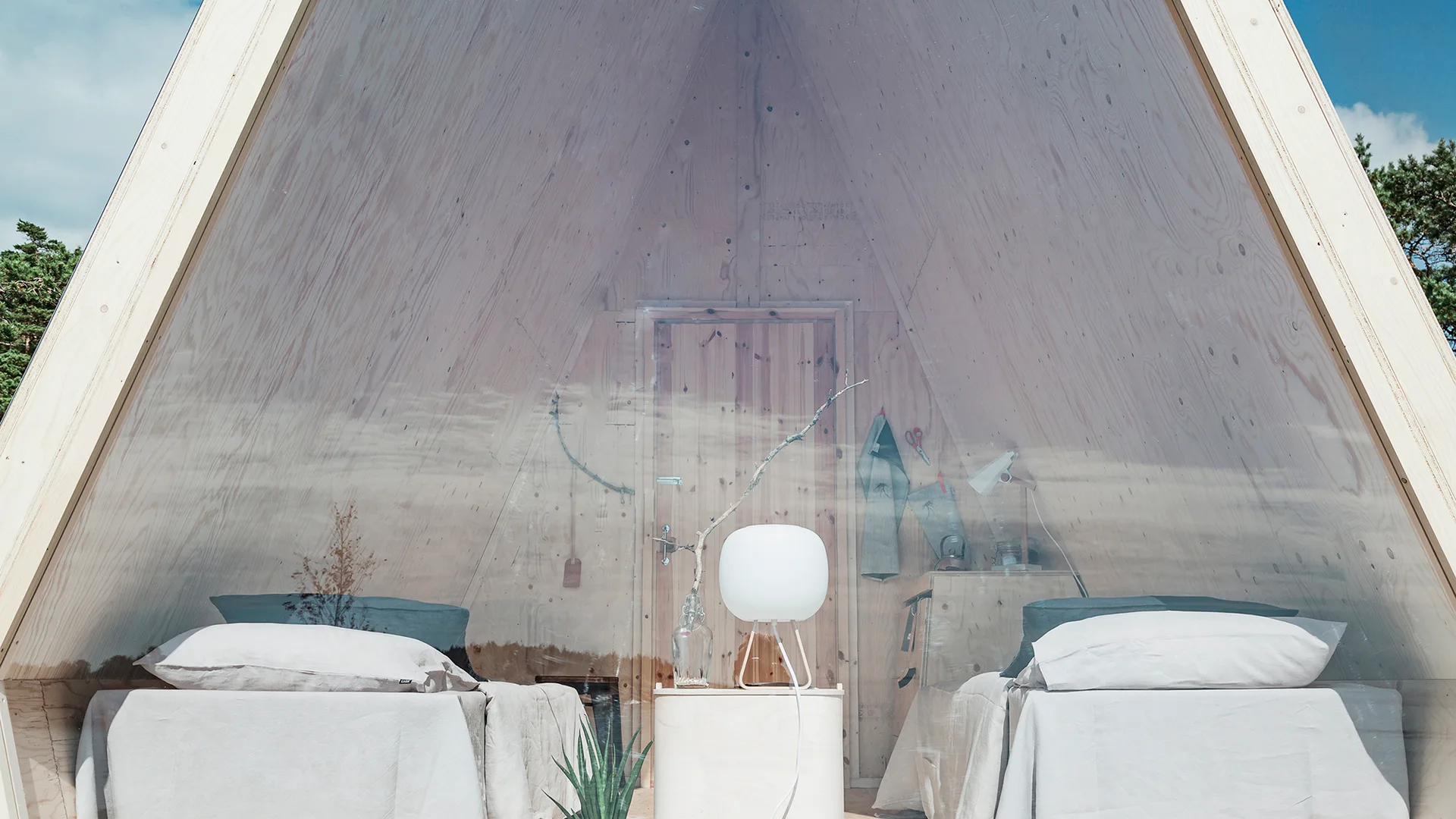 Two beds with light grey linens sit symmetrically inside a small A-frame cabin with light plywood walls and a glass front.