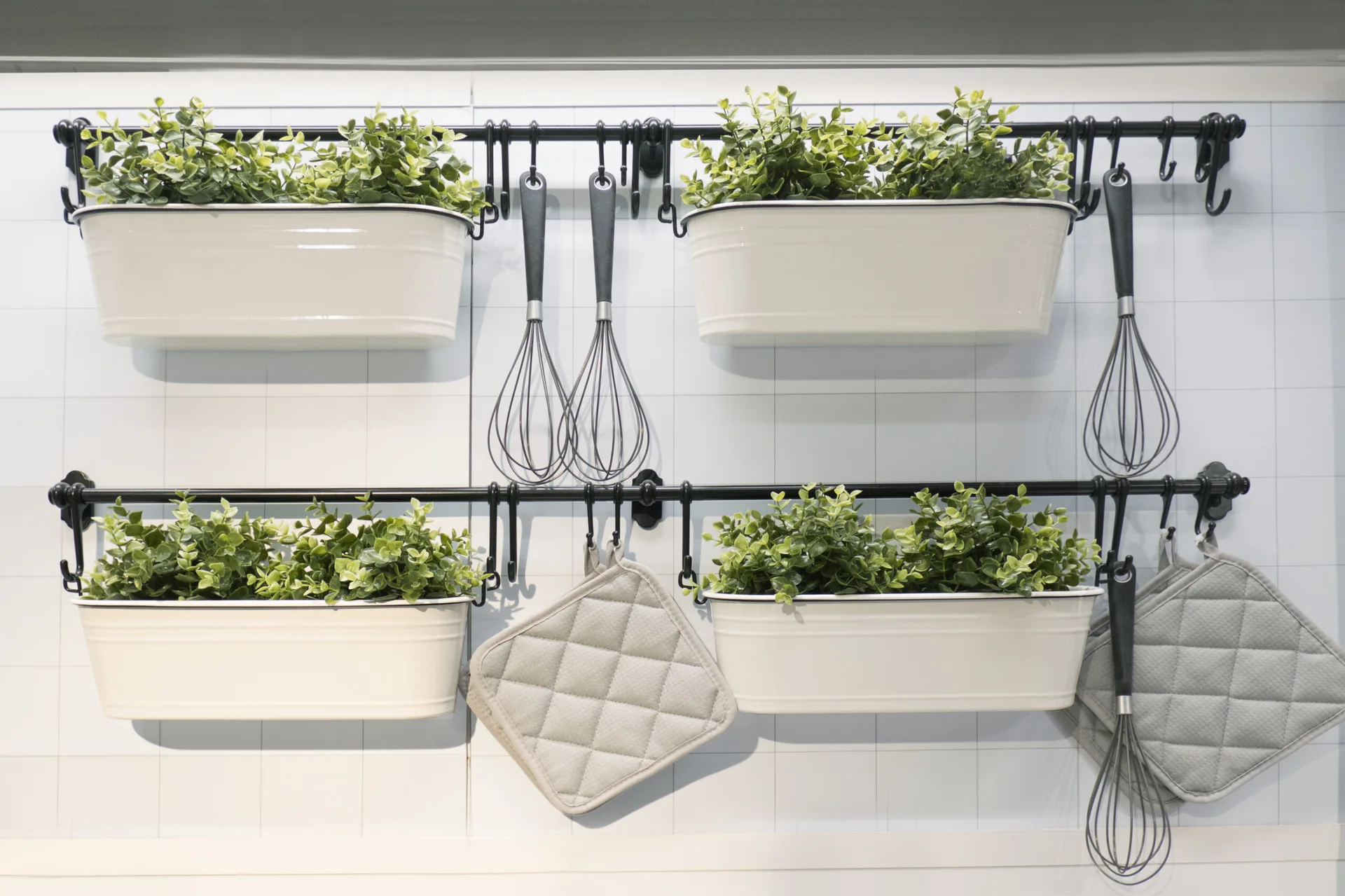 Black metal rails mounted on a white grid-tiled wall hang four white rectangular planters with green plants, silver whisks, and grey potholders.