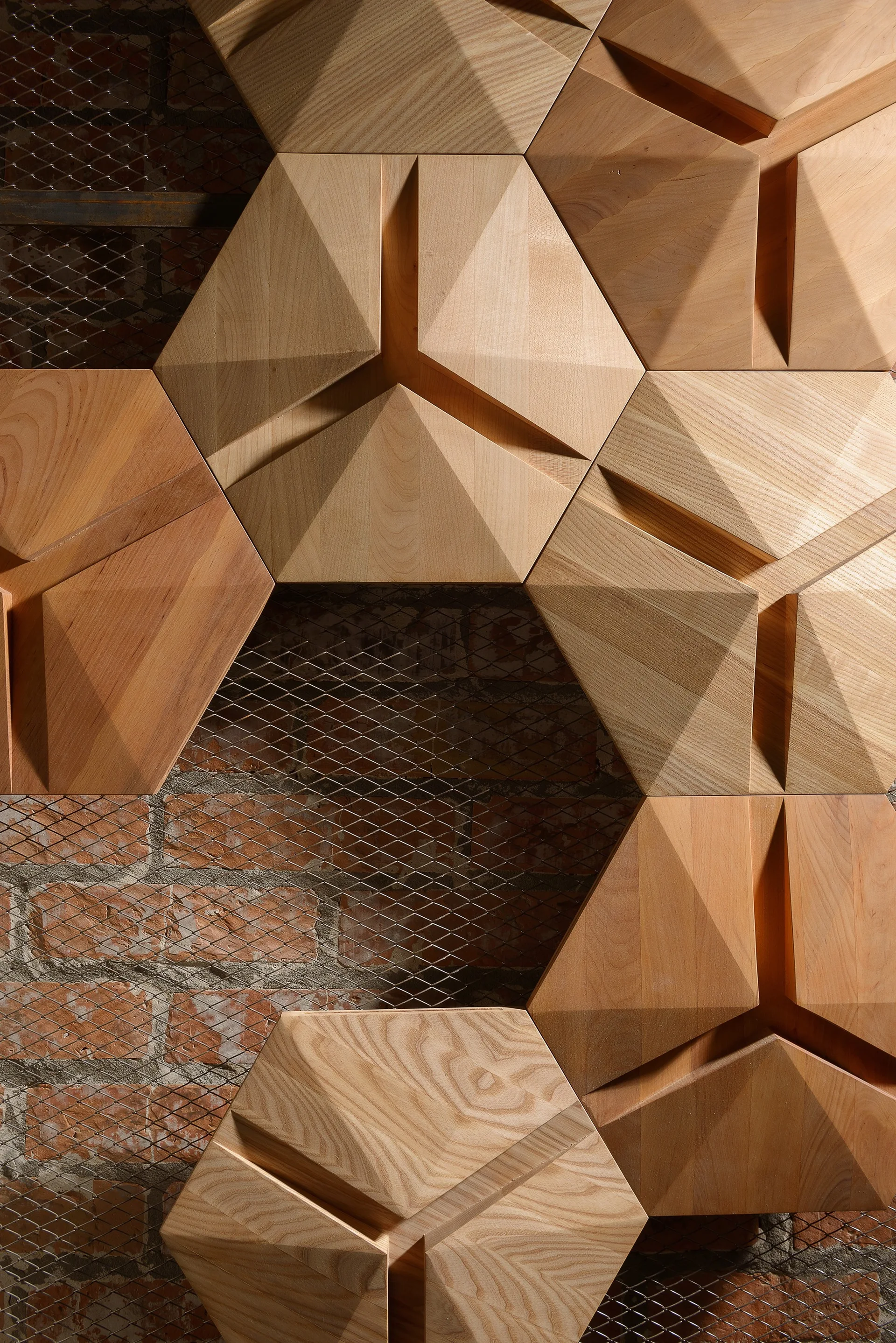 Modular faceted wooden wall tiles arranged over a layer of metal wire mesh and exposed red brick.
