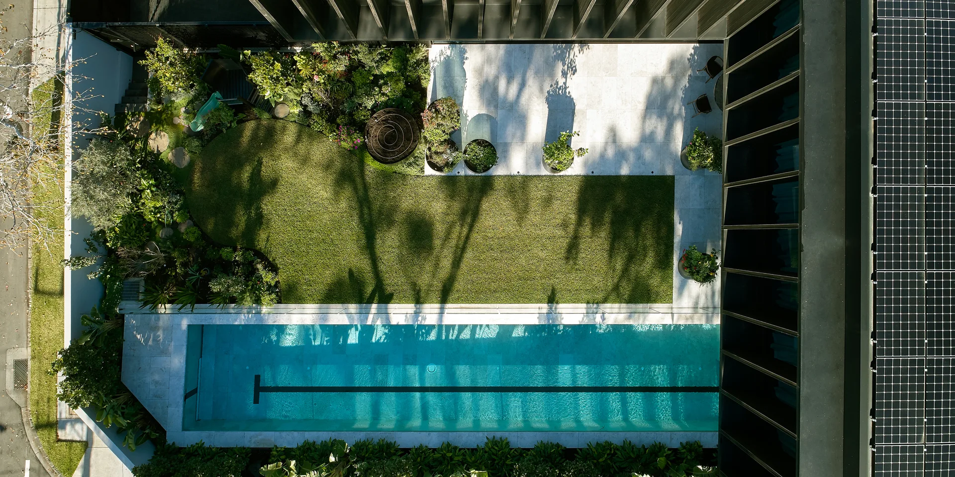 An aerial view of a modern backyard featuring a long blue lap pool, a rectangular green lawn, a tiled patio, and solar panels on the roof.
