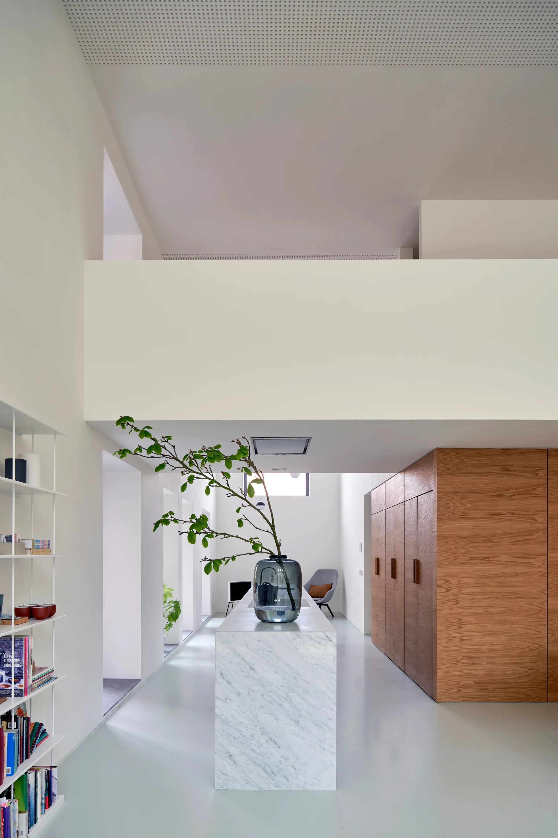 A minimalist kitchen features a central white marble island with a leafy branch in a vase, set against tall wooden cabinets and clean white walls.