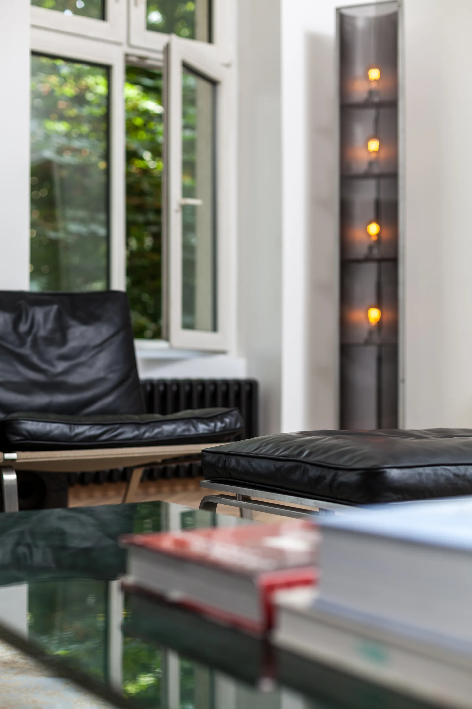 A modern living room features a black leather armchair and ottoman set next to a tall, glowing vertical tower floor lamp and a large window.