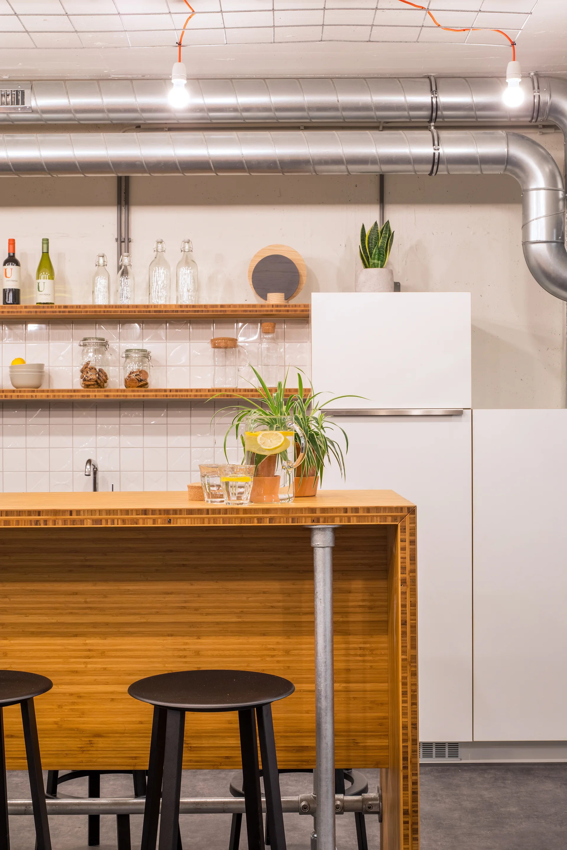 A modern industrial kitchen features a wood-grain island with black bar stools, a white tiled backsplash, and exposed silver ductwork on the ceiling.