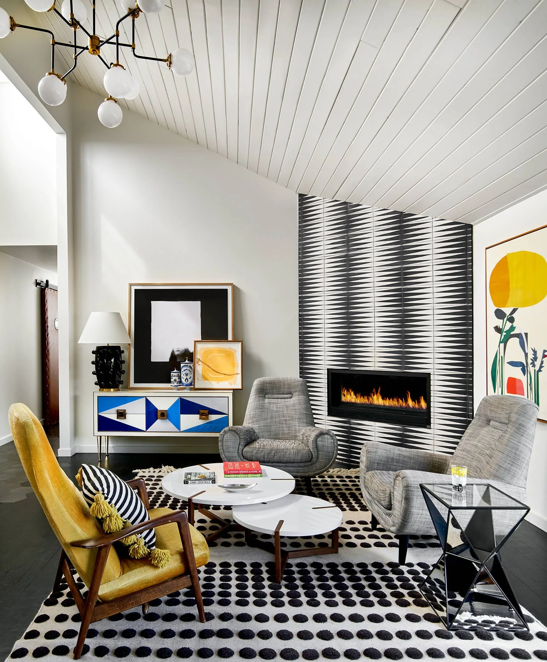 A modern living room features a fireplace with black and white chevron tiles, gray armchairs, a yellow chair, and a white planked vaulted ceiling.