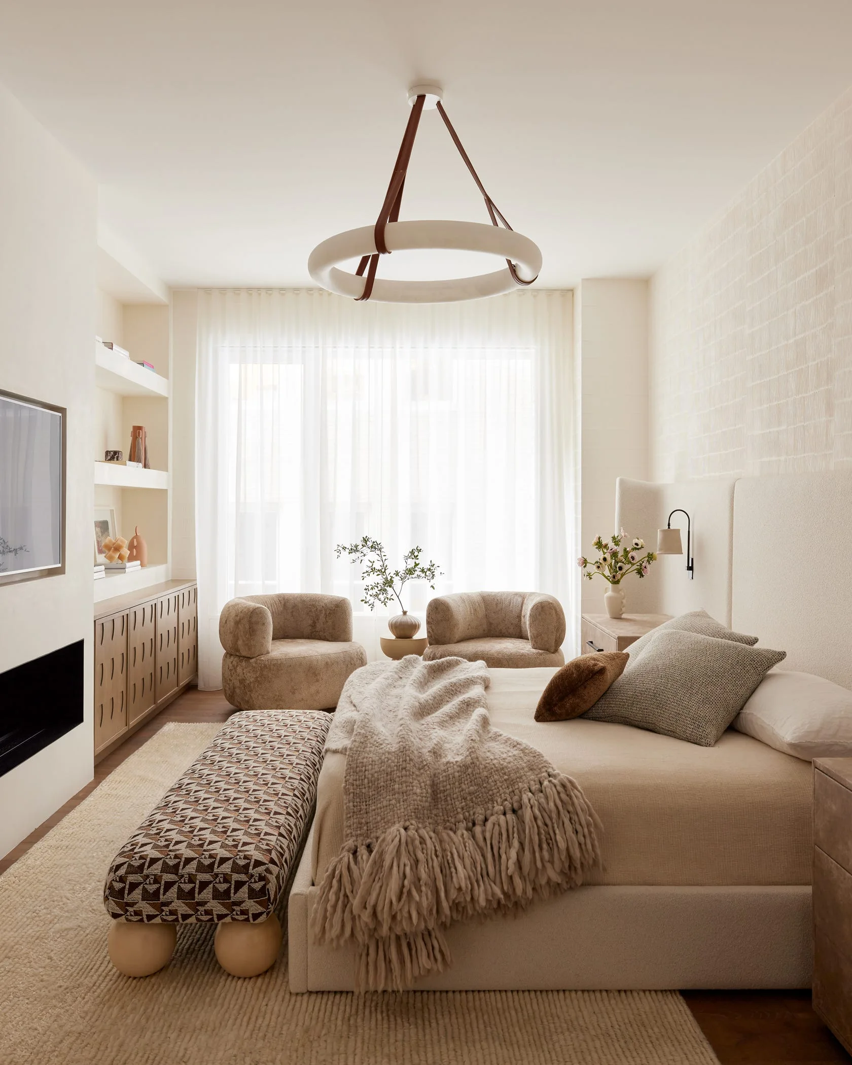 A cream-colored bedroom features an upholstered bed, a patterned bench, a circular chandelier, and two curved armchairs by a large window.