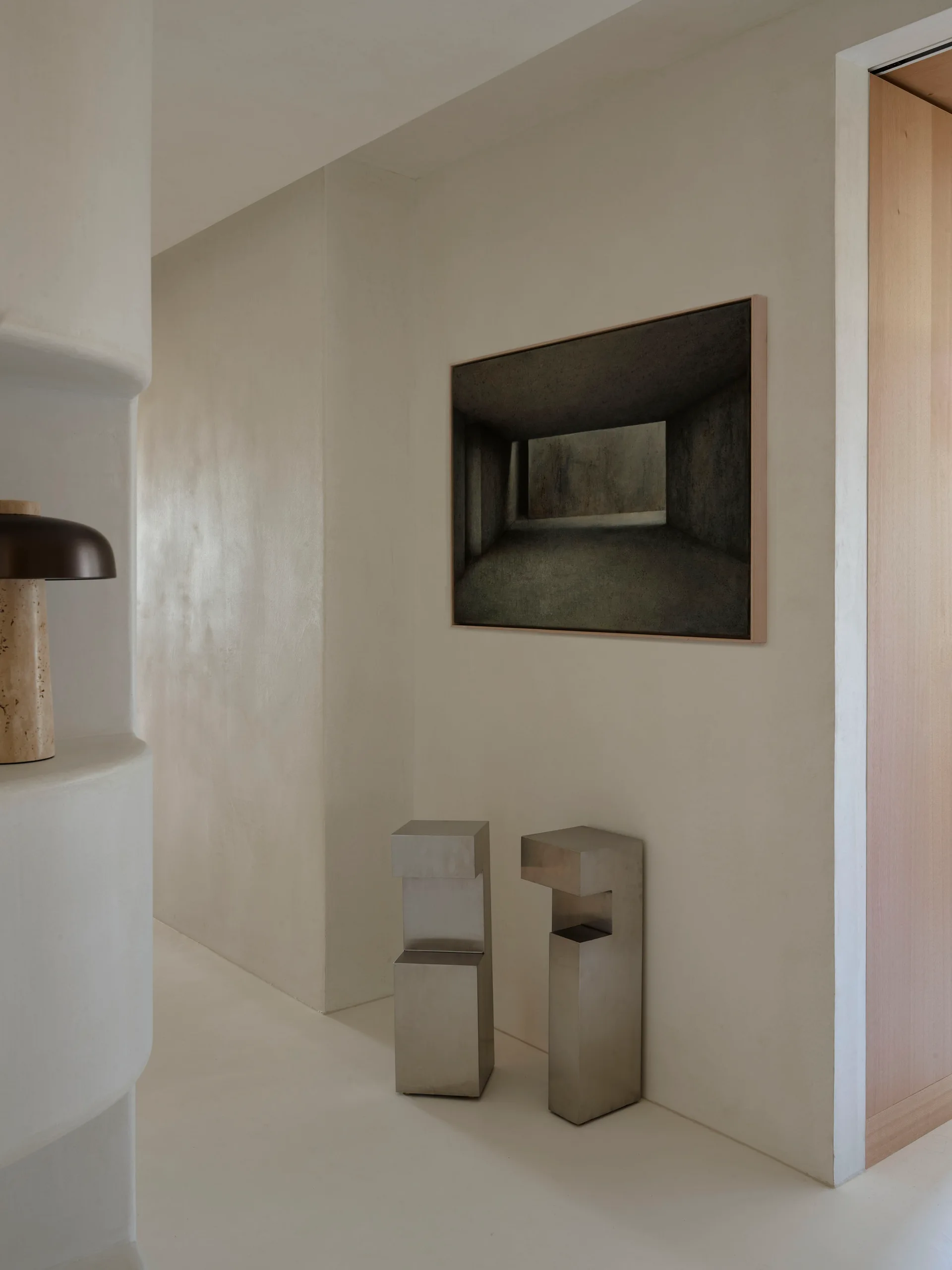 A minimalist cream hallway features curved plaster walls, a framed painting of a corridor, and two geometric metal sculptures on the floor.
