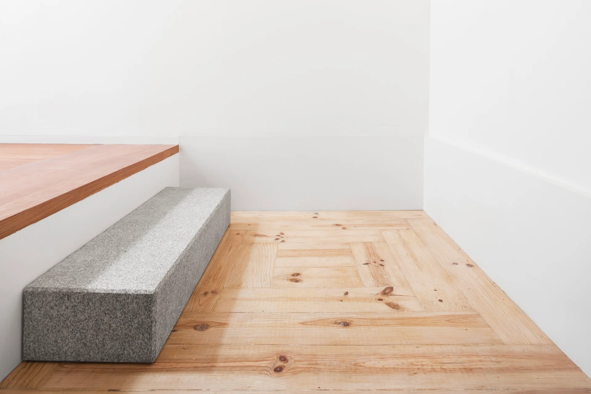 A minimalist interior detail showing knotty wood parquet flooring meeting a grey granite block step and a plain white wall.