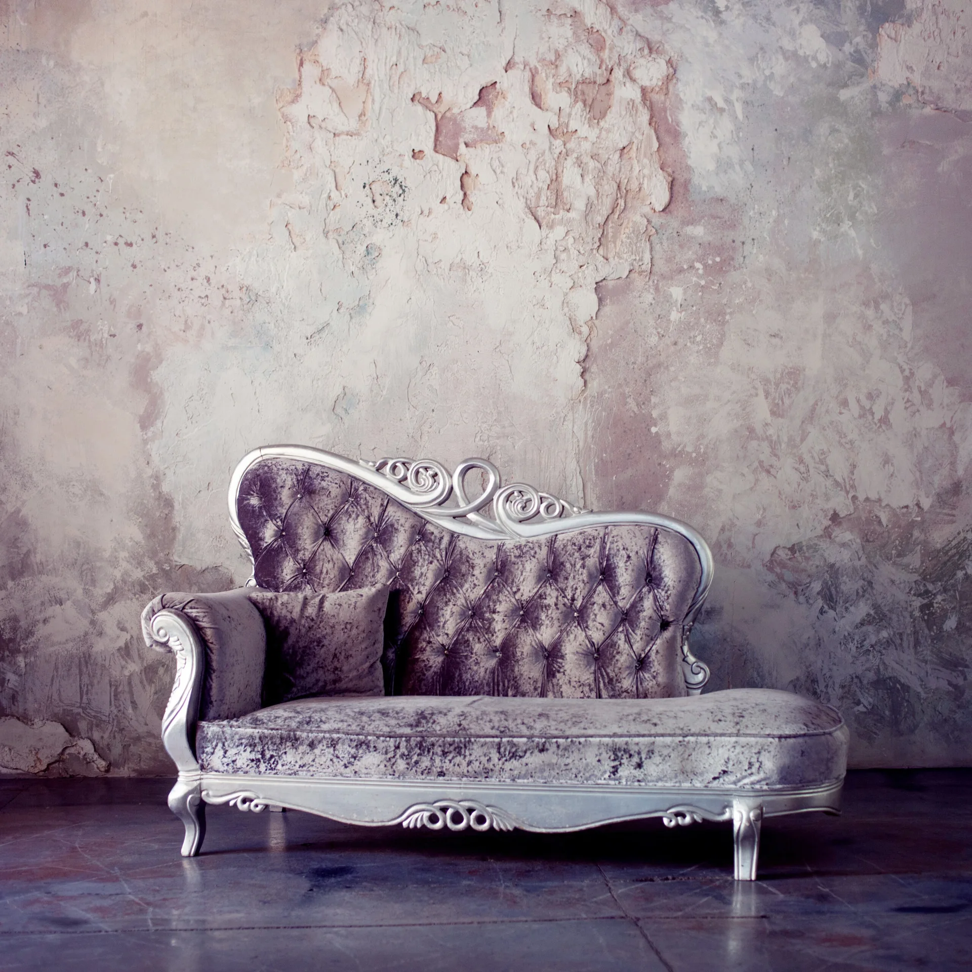 A tufted silver velvet chaise longue sits on a grey concrete floor against a weathered, peeling plaster wall.