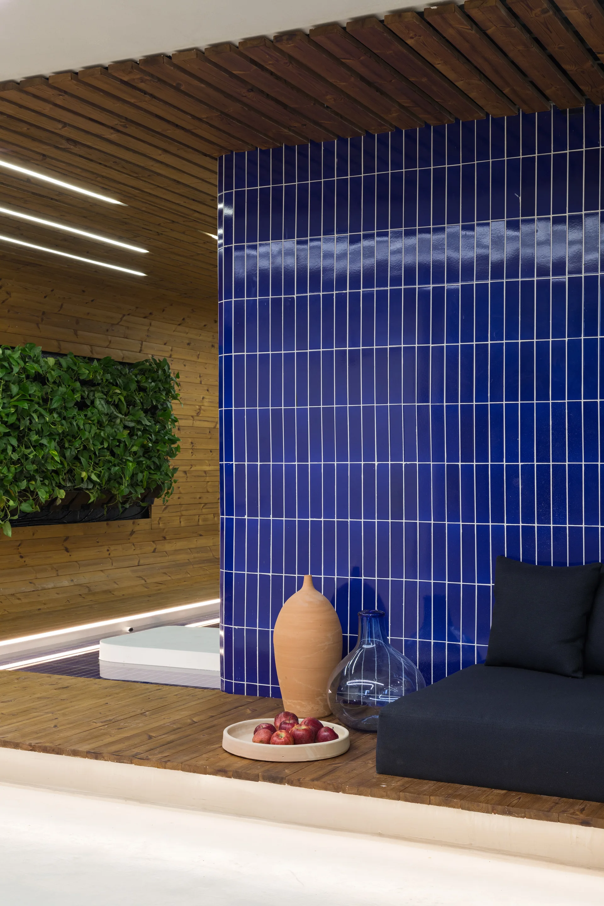 An indoor lounge features a glossy cobalt blue tiled wall, black platform seating, a slatted wood ceiling, and decorative earthenware vases.