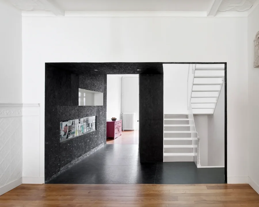 A black textured wood architectural block creates an entryway corridor between a white room with wood floors and a staircase.