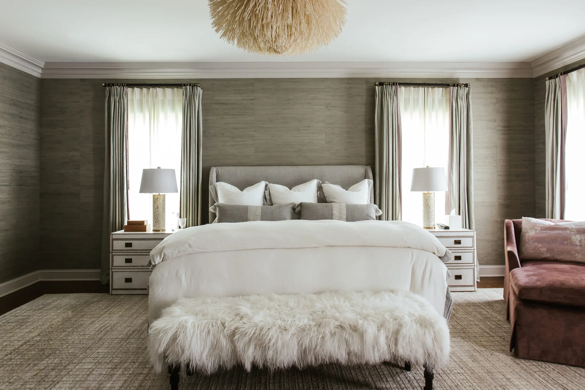 A symmetrical bedroom with a grey upholstered bed, textured grasscloth wallpaper, two white nightstands with lamps, and a furry bench.