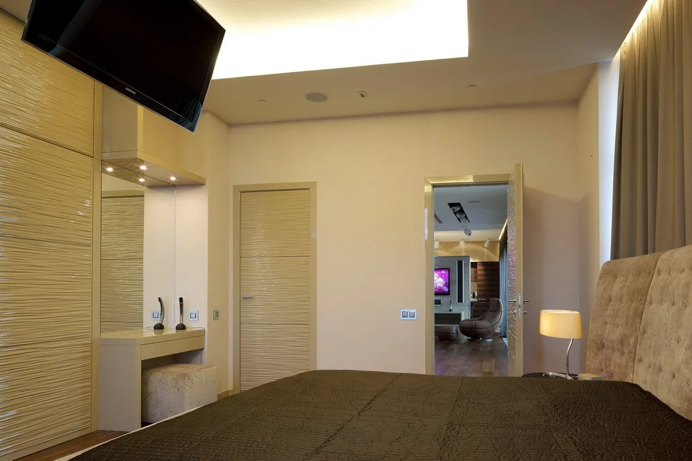 A modern bedroom featuring a tufted headboard, a built-in vanity, textured wardrobe doors, and glowing ceiling cove lighting.