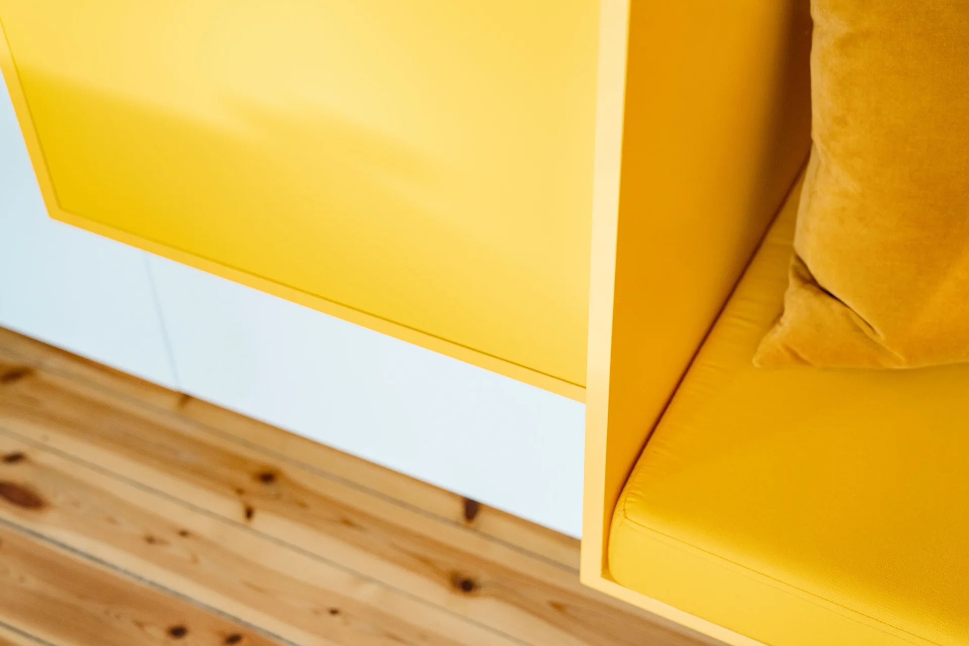 A close-up of a bright yellow built-in bench with a matching cushion and pillow set against white paneling and a light wood floor.