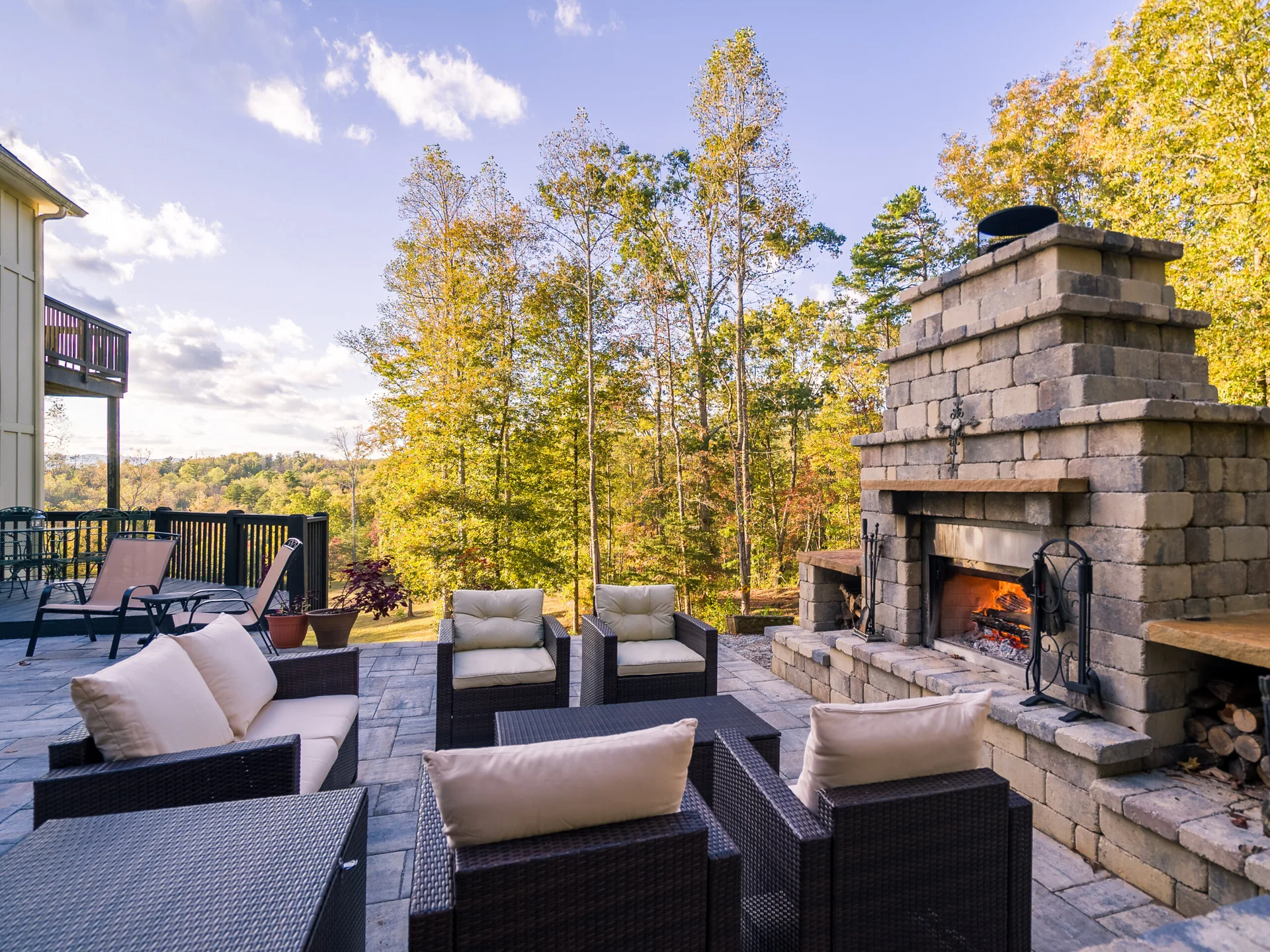 An outdoor stone patio features a large stone fireplace with a burning fire, surrounded by dark wicker armchairs with beige cushions.