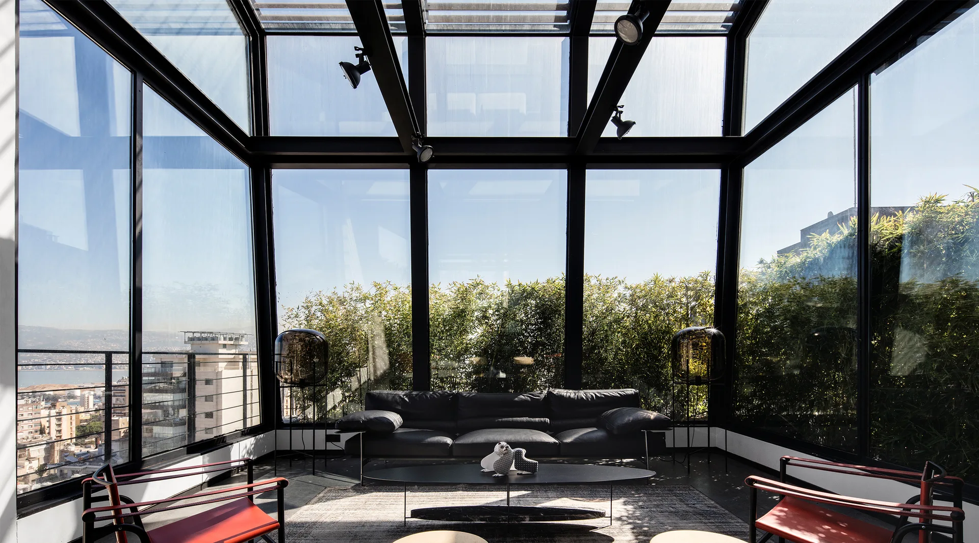 A modern glass-enclosed living room features a black leather sofa, red armchairs, a low coffee table, and views of greenery through the glass walls.