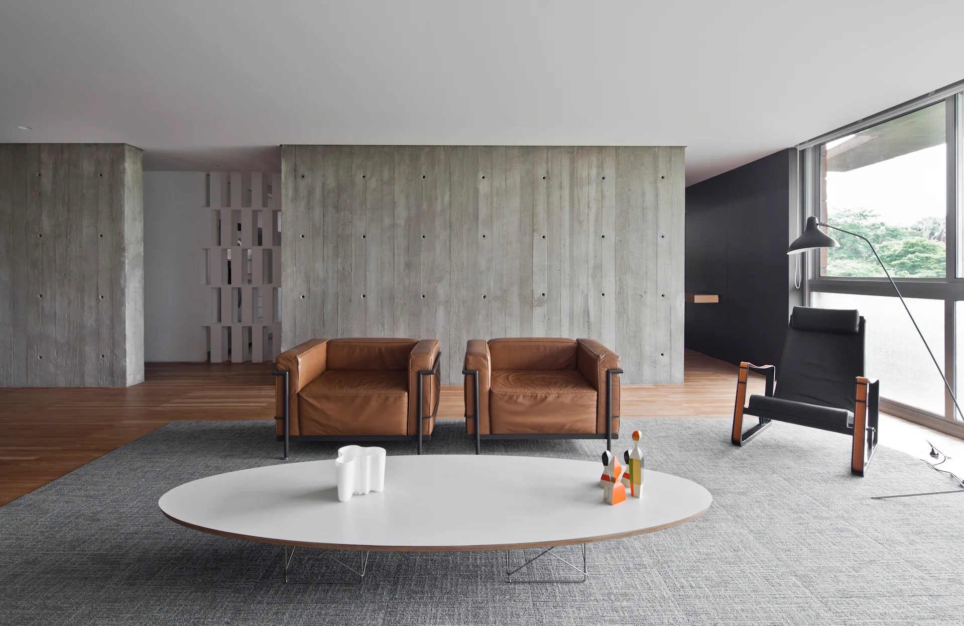 A minimalist living room features two tan leather armchairs, a white oval coffee table, and an exposed concrete wall.