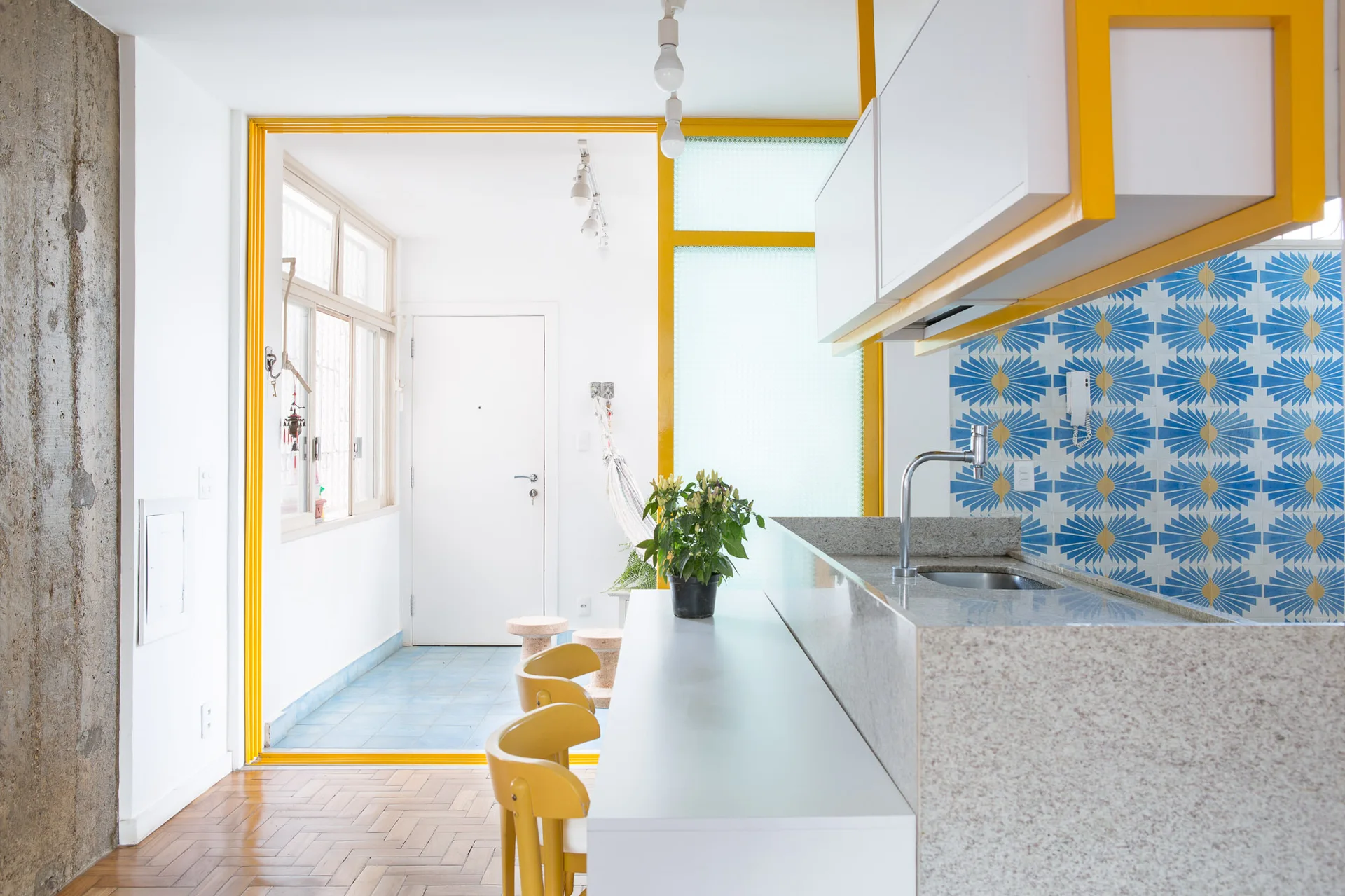 A modern kitchen features a stone-topped island with yellow bar stools, a blue patterned tile backsplash, and a bright yellow door frame.