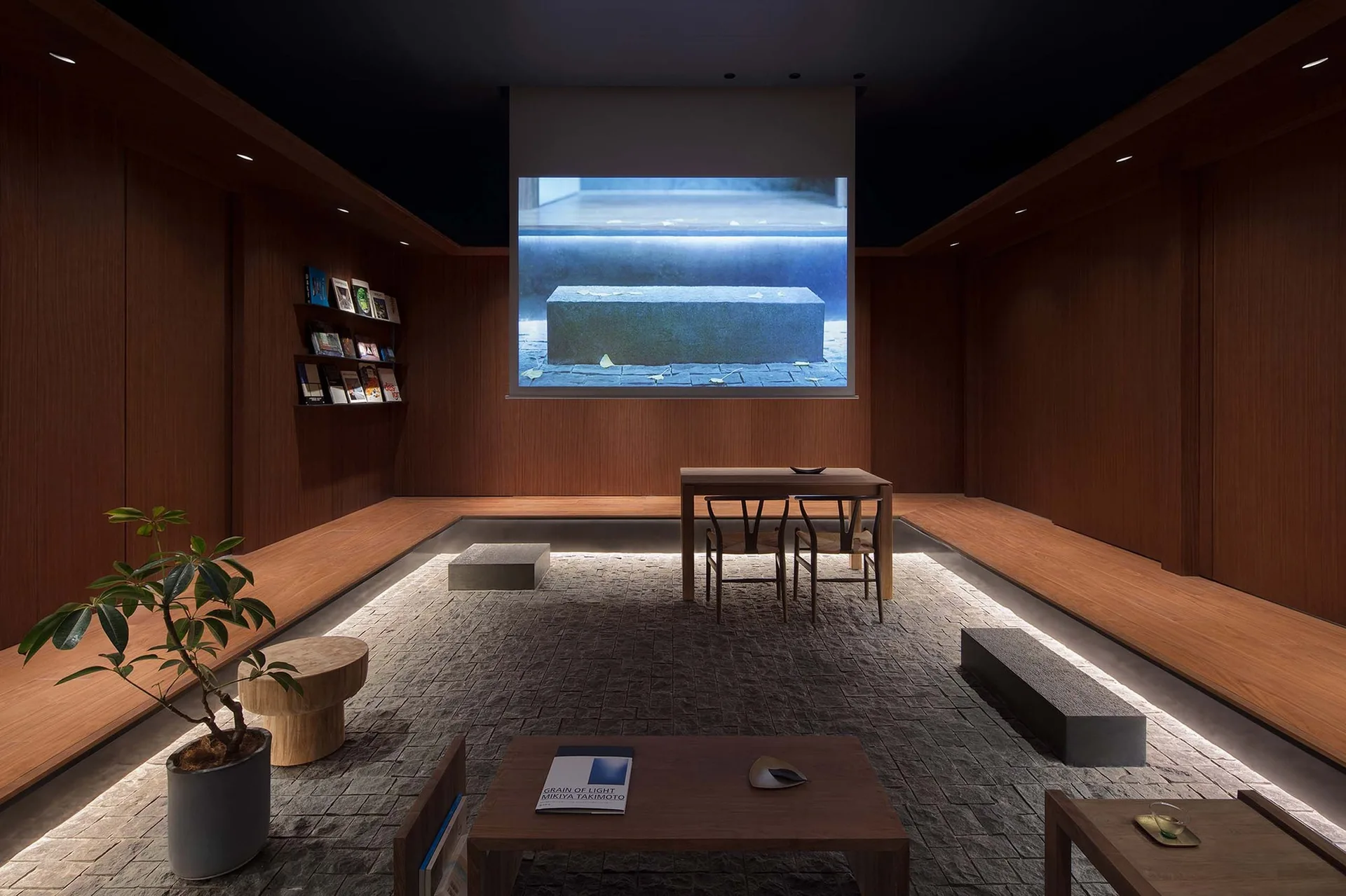 A minimalist sunken lounge features warm wood-paneled walls, grey cobblestone flooring with under-bench cove lighting, and a central projector screen.