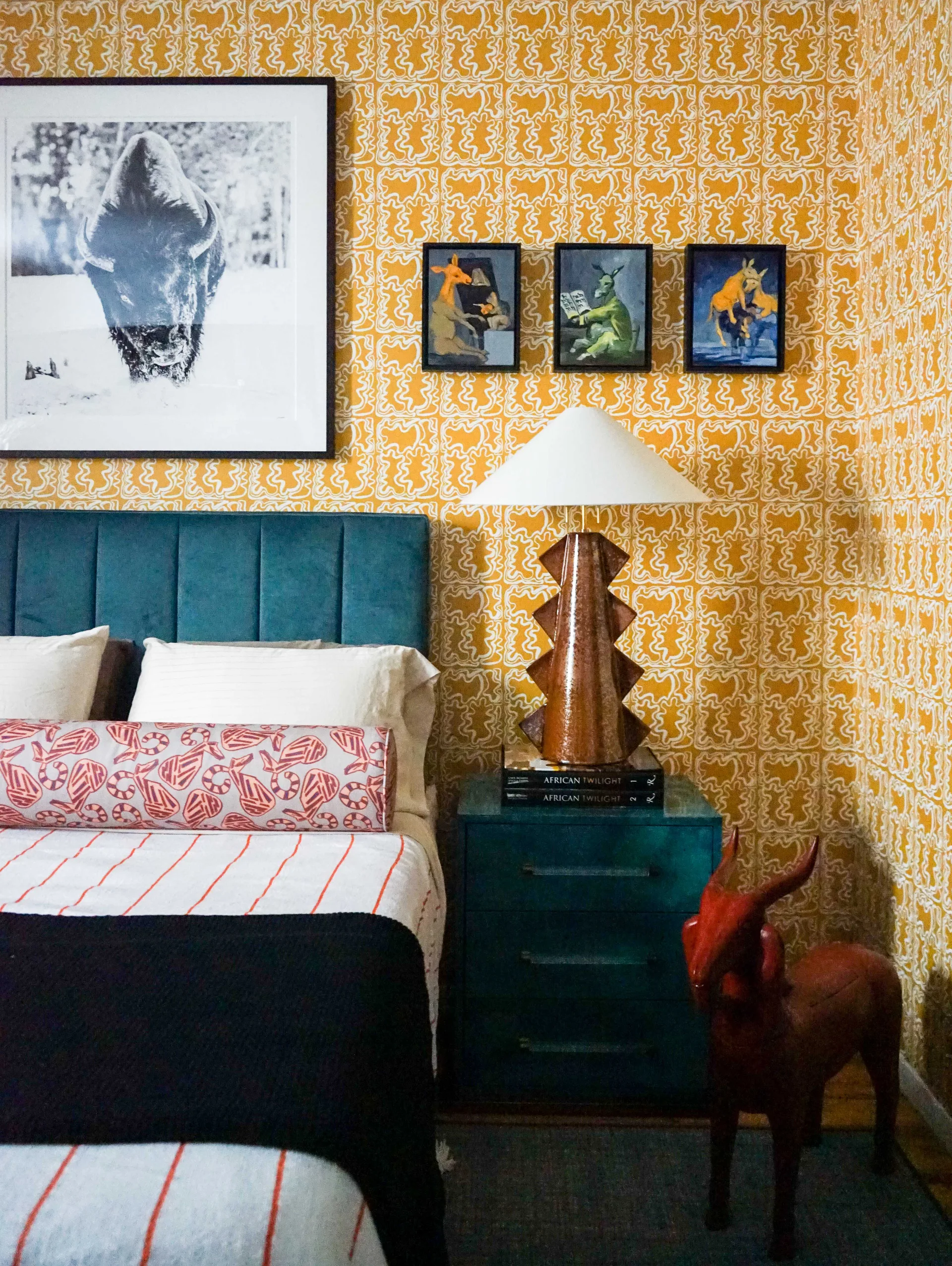 A maximalist bedroom corner featuring yellow patterned wallpaper, a teal channel-tufted headboard, and a teal nightstand with a sculpted lamp.