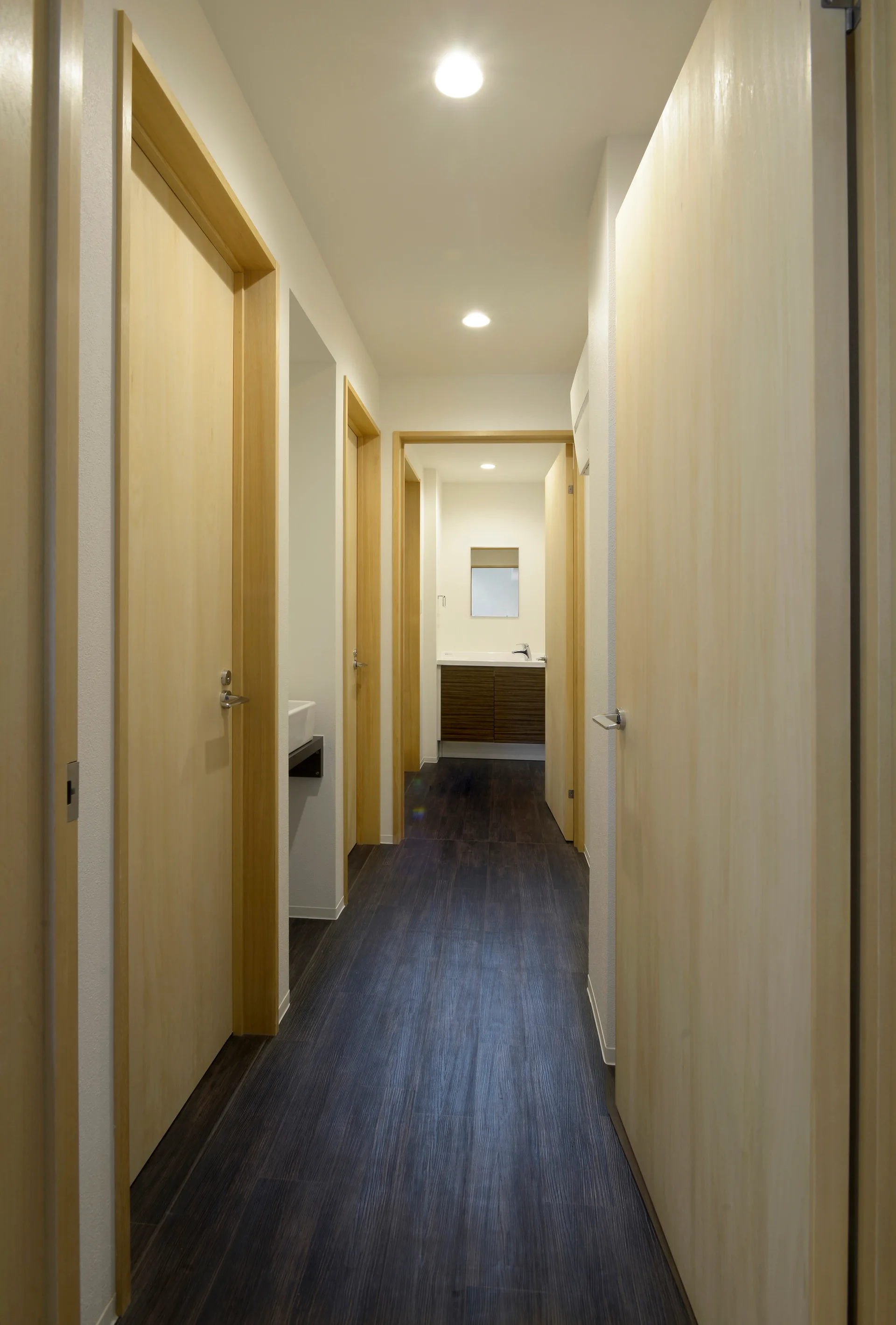 A narrow hallway with multiple light wood doors, white walls, and a dark wood floor leading to a bathroom vanity.