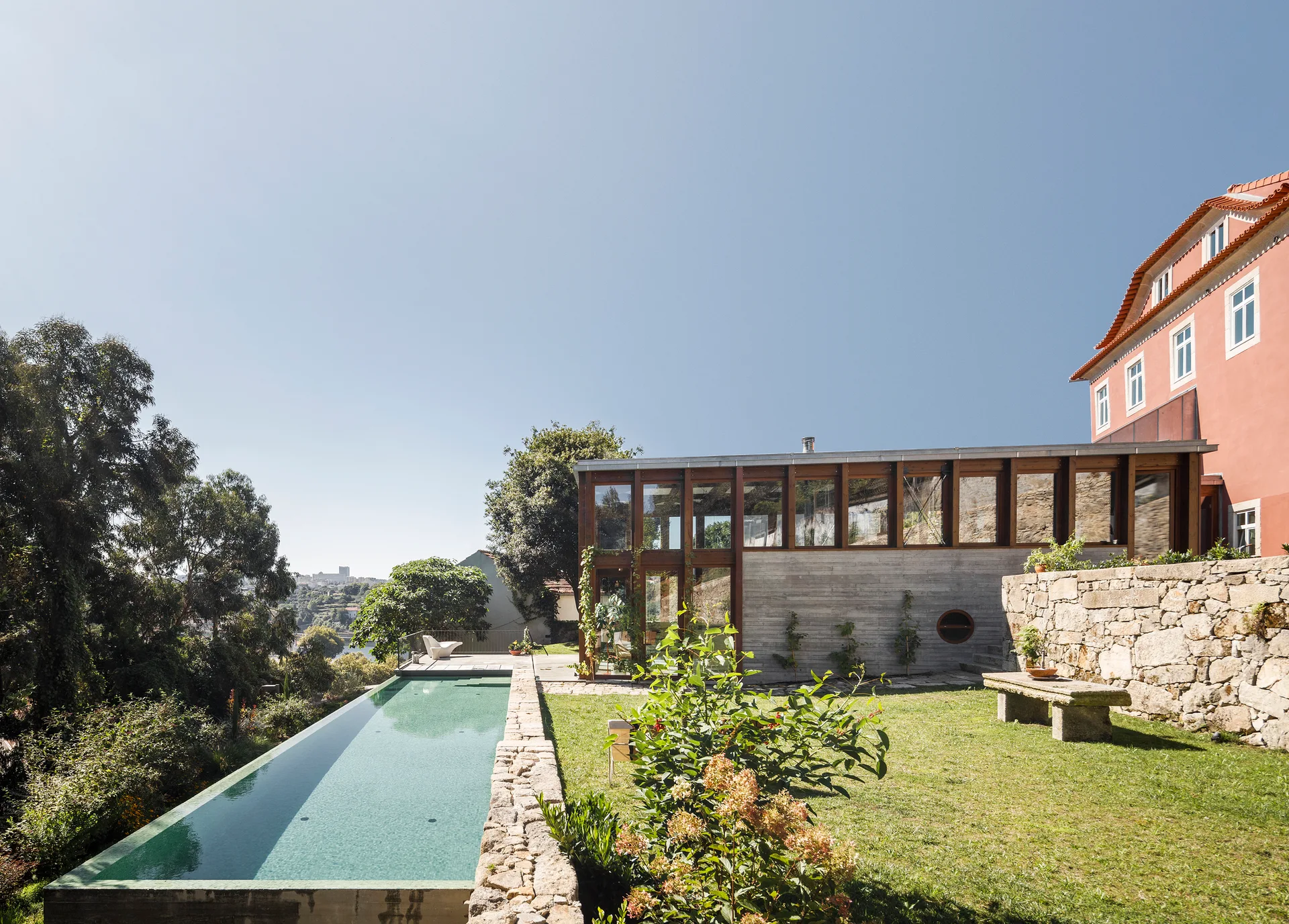 A long rectangular lap pool bordered by rustic stone masonry sits next to a green lawn and a modern glass and concrete house extension.