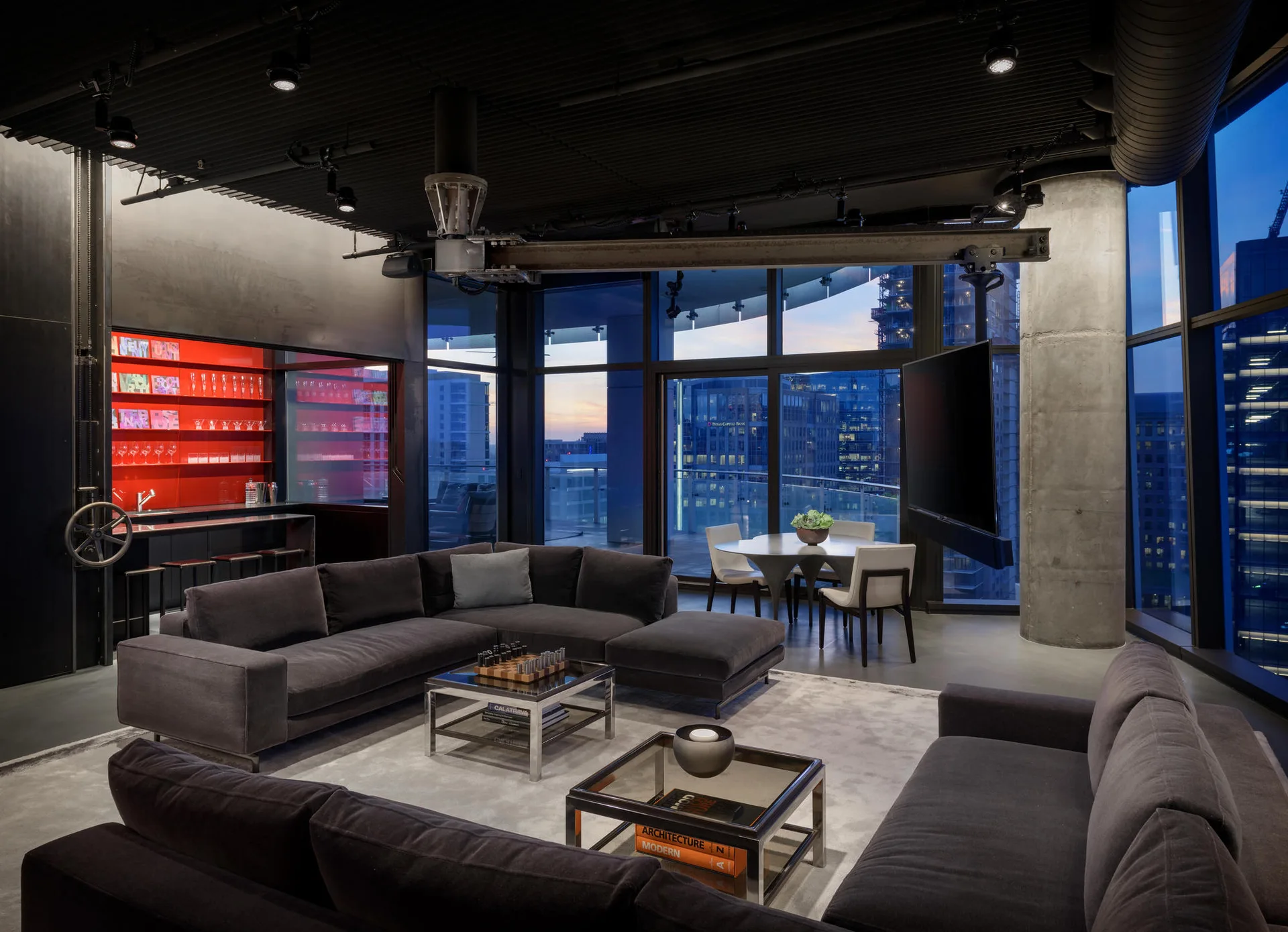 A modern industrial living room features grey sectional sofas, glass coffee tables, a concrete column, and floor-to-ceiling windows showing a city view.