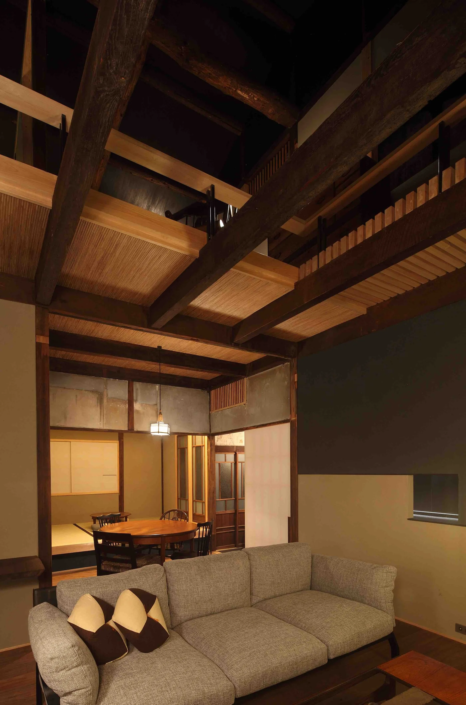 A grey upholstered sofa sits beneath towering dark wood ceiling beams in an open living space with a traditional Japanese dining room in the background.