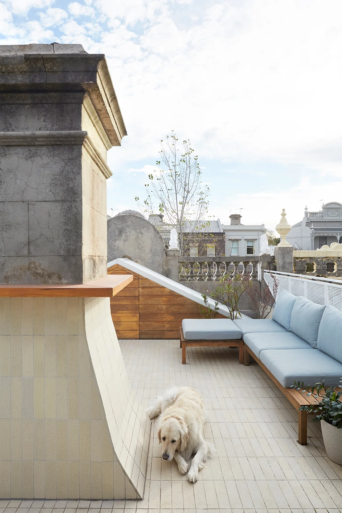 A light blue sectional sofa sits on a tiled rooftop terrace alongside a large chimney structure, a stone balustrade, and a sleeping dog.