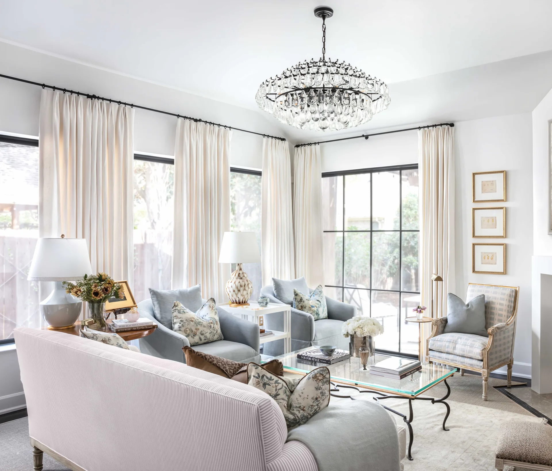 A bright living room features a pinstriped sofa, light blue armchairs, a glass coffee table, and a large tiered crystal chandelier.