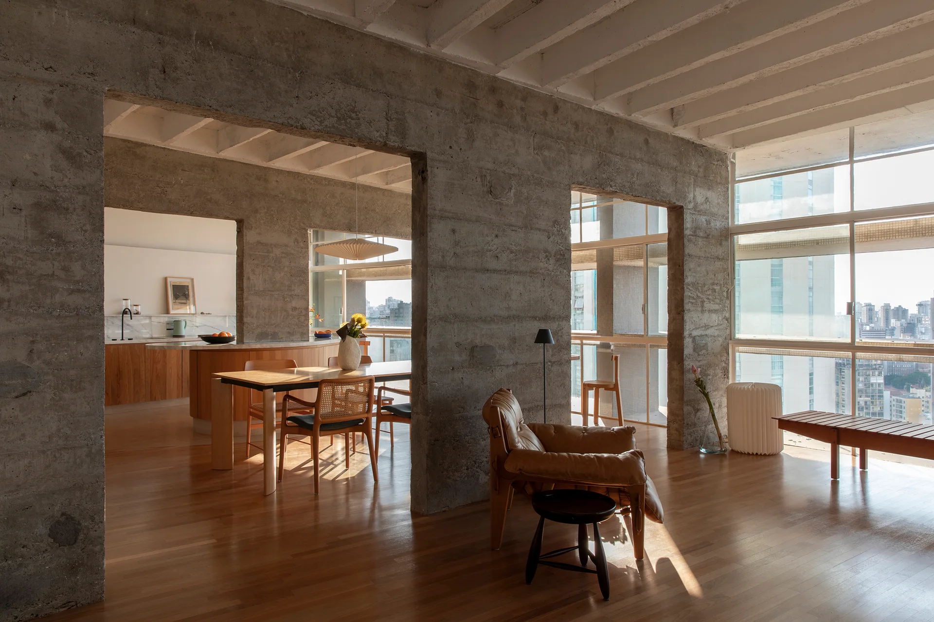 An open-concept apartment interior features raw concrete structural walls, warm wood flooring, a tan leather armchair, and a wooden dining table.